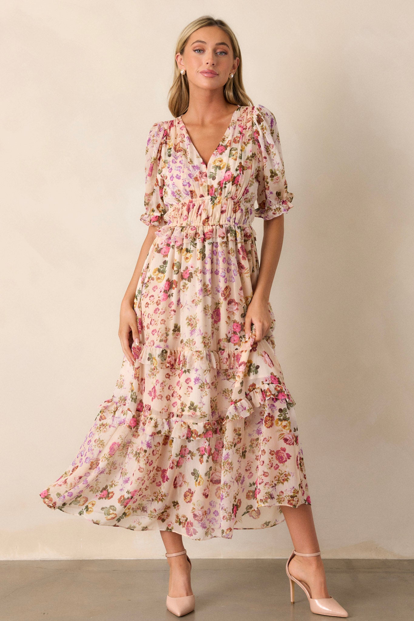 Front view of this beige dress that features a v-neckline, two thin elastic waist bands, a tiered design, ruffled trim on the tiers, a pink floral design, a midi length and half length puff sleeves with elastic cuffs.