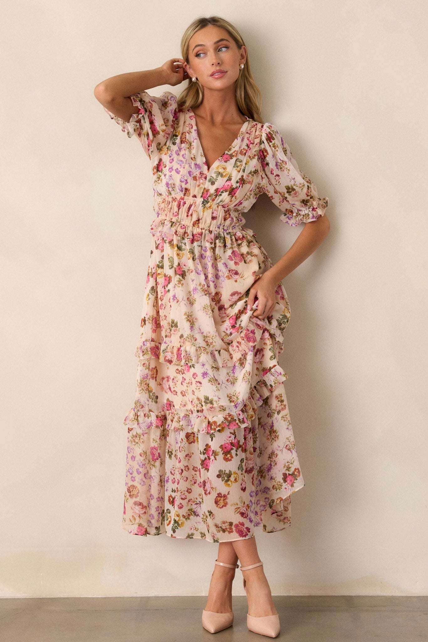 Full body view of this beige dress that features a v-neckline, two thin elastic waist bands, a tiered design, ruffled trim on the tiers, a pink floral design, a midi length and half length puff sleeves with elastic cuffs.