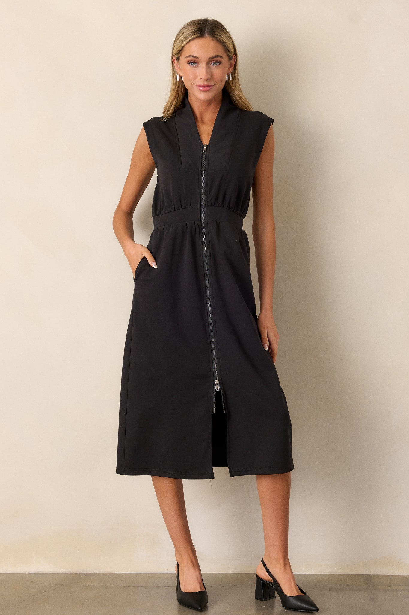 Full body view of this black dress that features a v-neckline, a slight collar, a zipper front, functional hip pockets, a middle split in the hem and a sleeveless design.