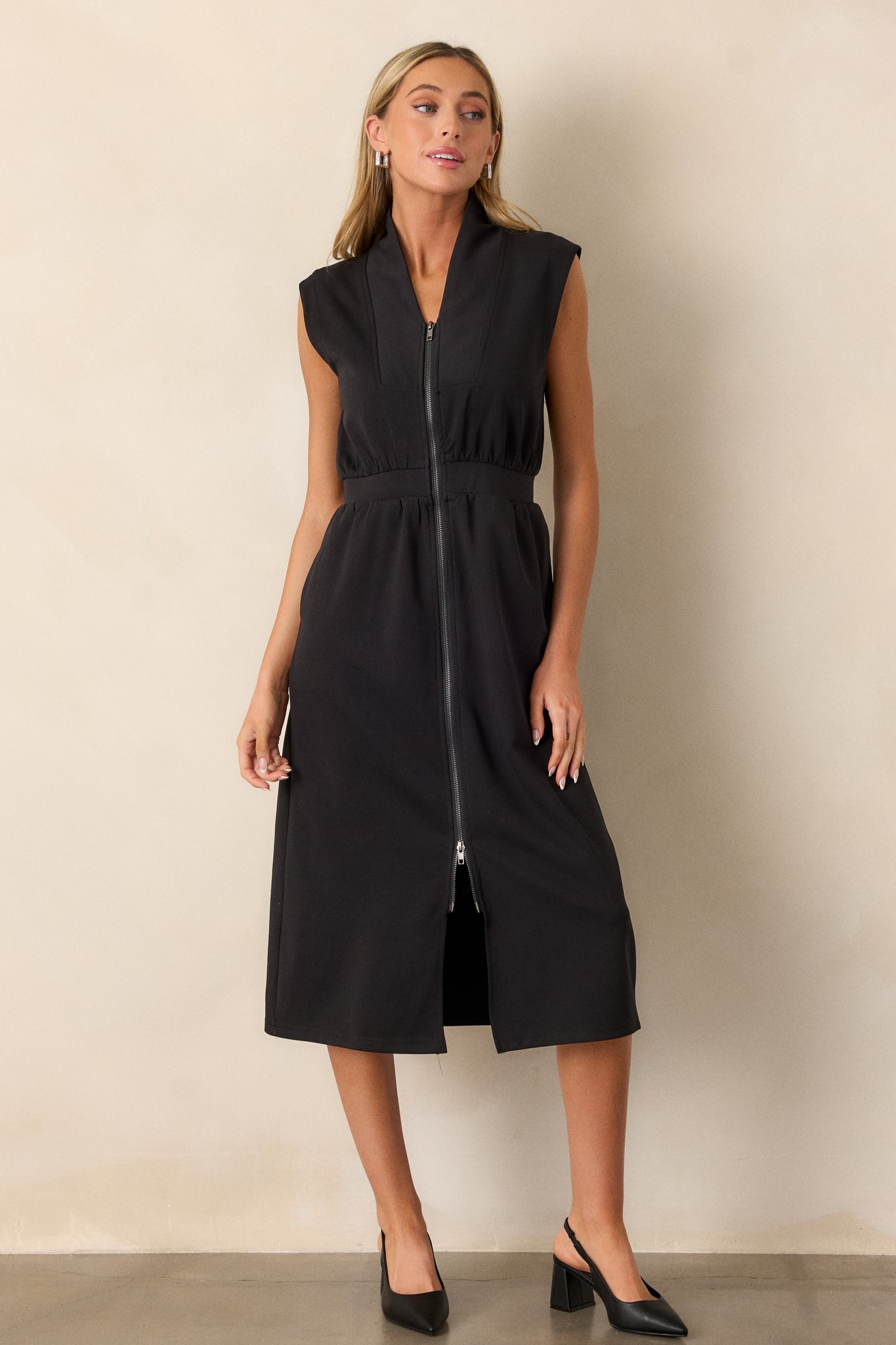 Angled view of this black dress that features a v-neckline, a slight collar, a zipper front, functional hip pockets, a middle split in the hem and a sleeveless design.