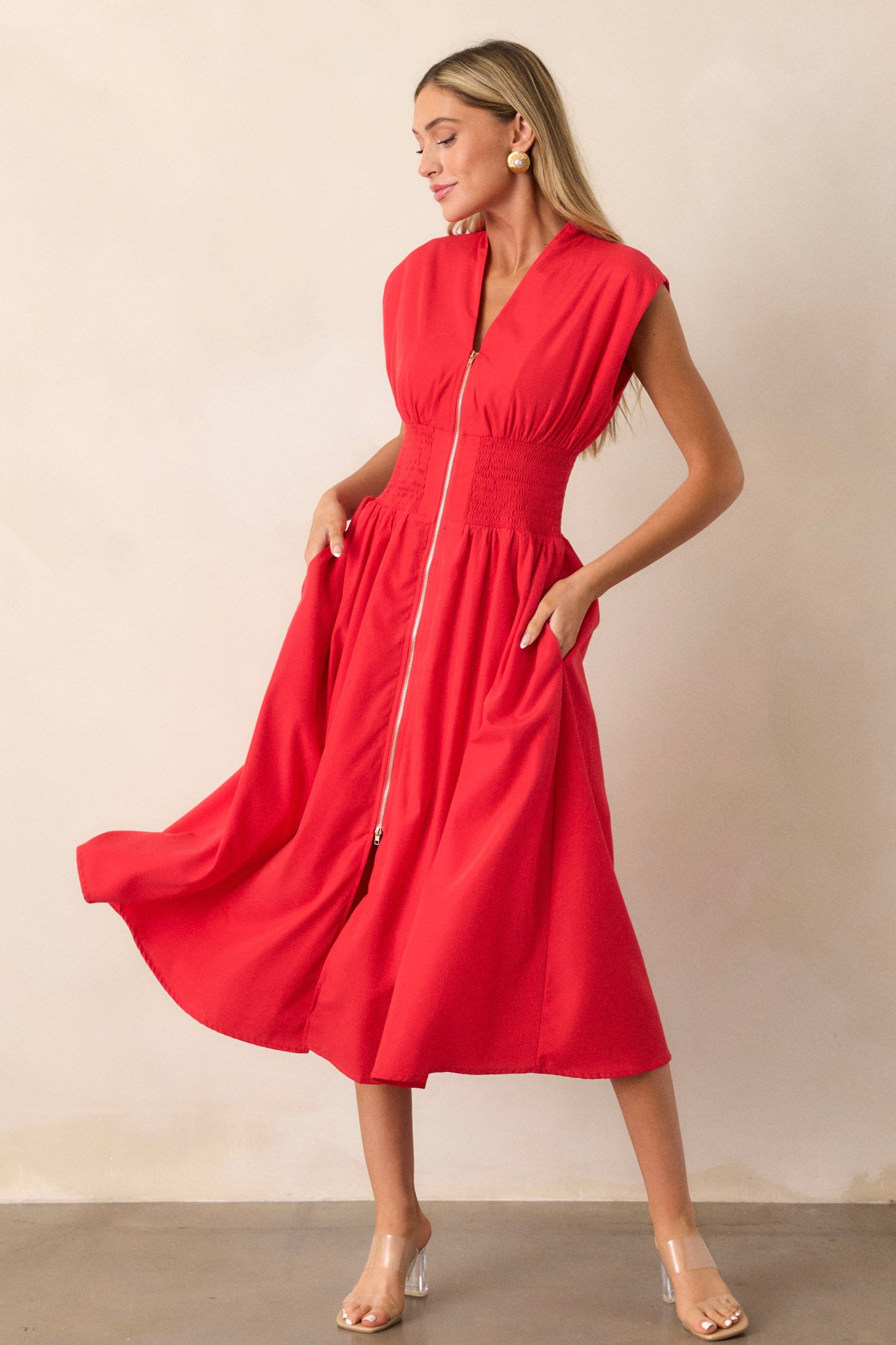 Angled full body view of this red midi dress that features a v-neckline, padded shoulders, a full zipper front, a fully smocked waist, functional hip pockets, and a front slit.