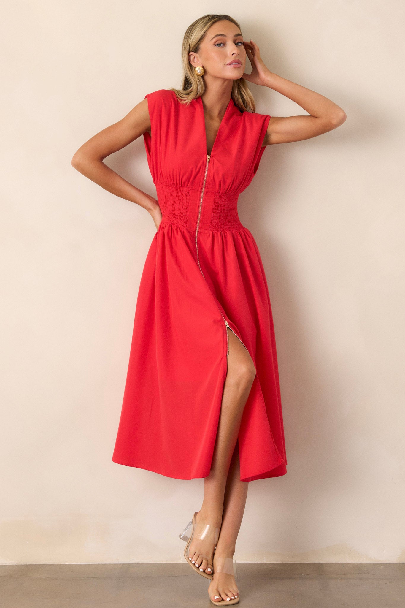 Angled front view of this red midi dress that features a v-neckline, padded shoulders, a full zipper front, a fully smocked waist, functional hip pockets, and a front slit.