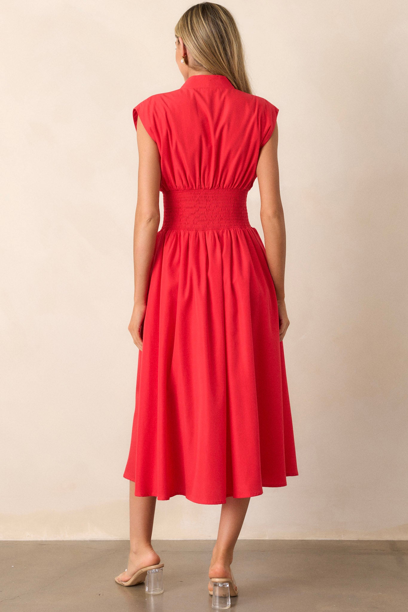 Back view of this red midi dress that features a v-neckline, padded shoulders, a full zipper front, a fully smocked waist, functional hip pockets, and a front slit.