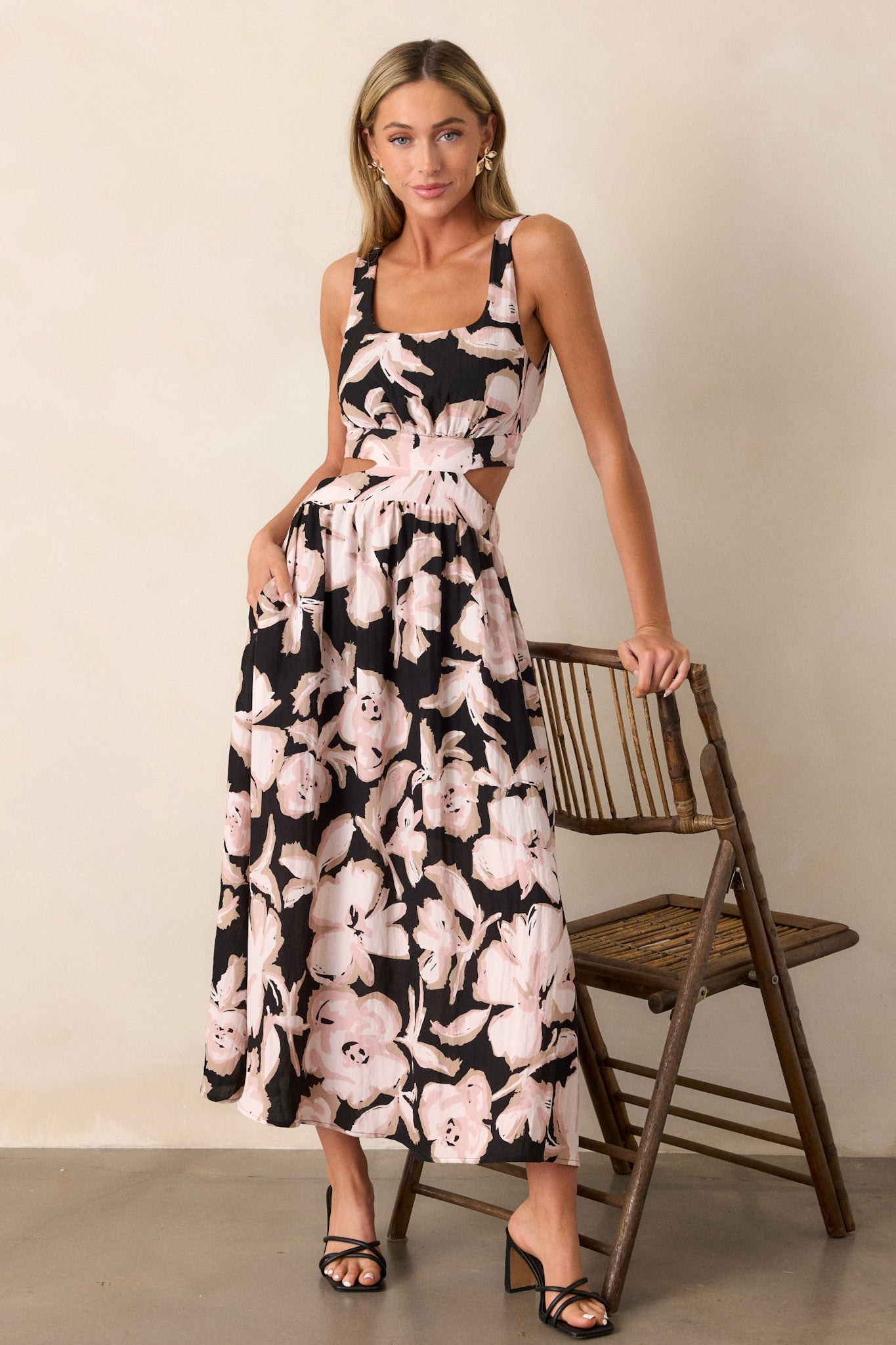 Full body view of this black maxi dress that features a square neckline, thick straps, an elastic back insert, a self-tie back feature, waist cutouts with a smocked back insert, functional hip pockets, and an all over floral pattern.