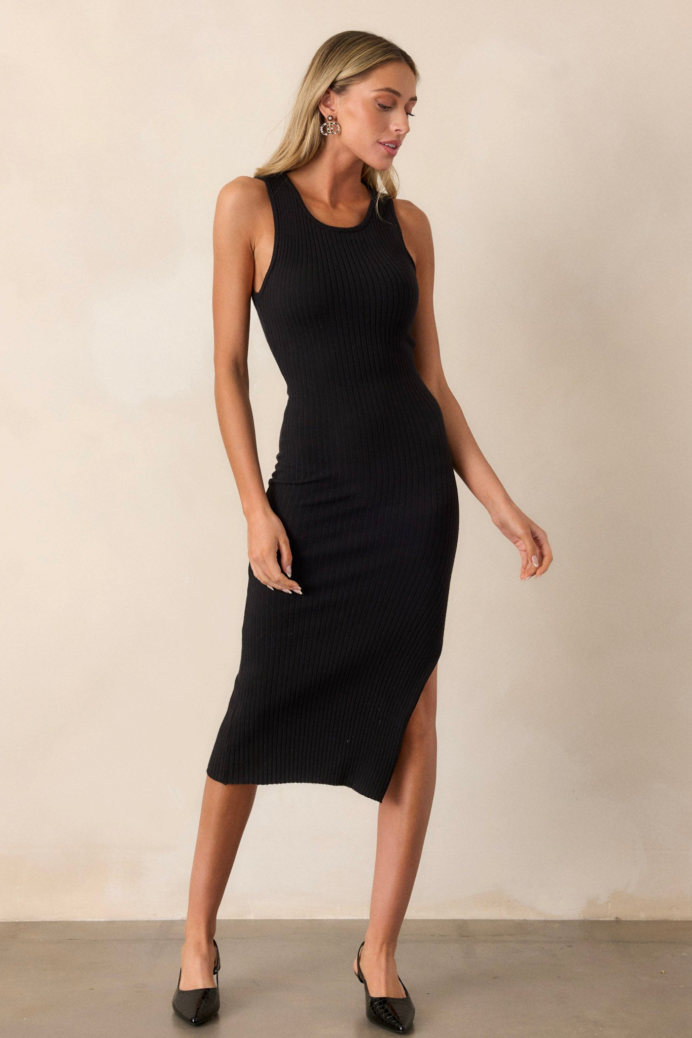 Full body view of this black bodycon dress featuring a high crew neckline, a ribbed material throughout, a slit up the leg and a sleeveless design.