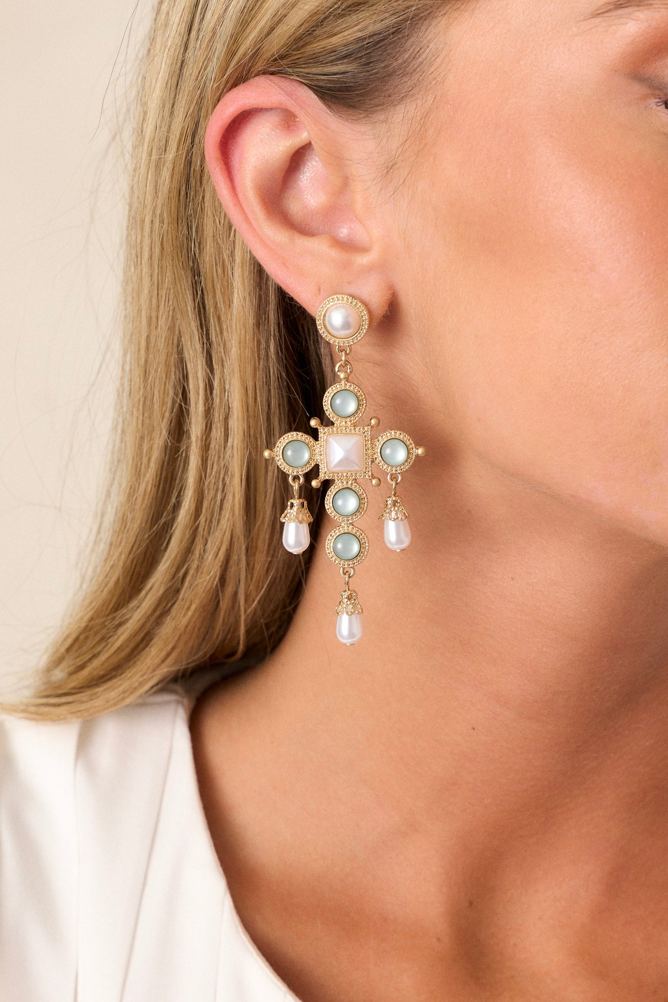 A close-up view of the gold earrings, featuring an ivory faux pearl stud at the top and a drop cross-shaped design with sage slightly iridescent faux stones.