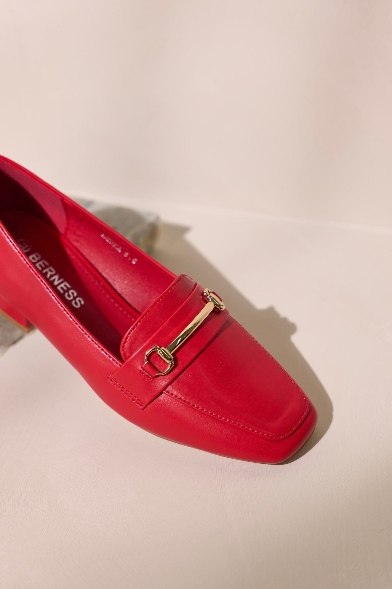 Close-up of the toe of these  red loafers that feature a slip on design, a small heel, a square toe, and gold hardware.