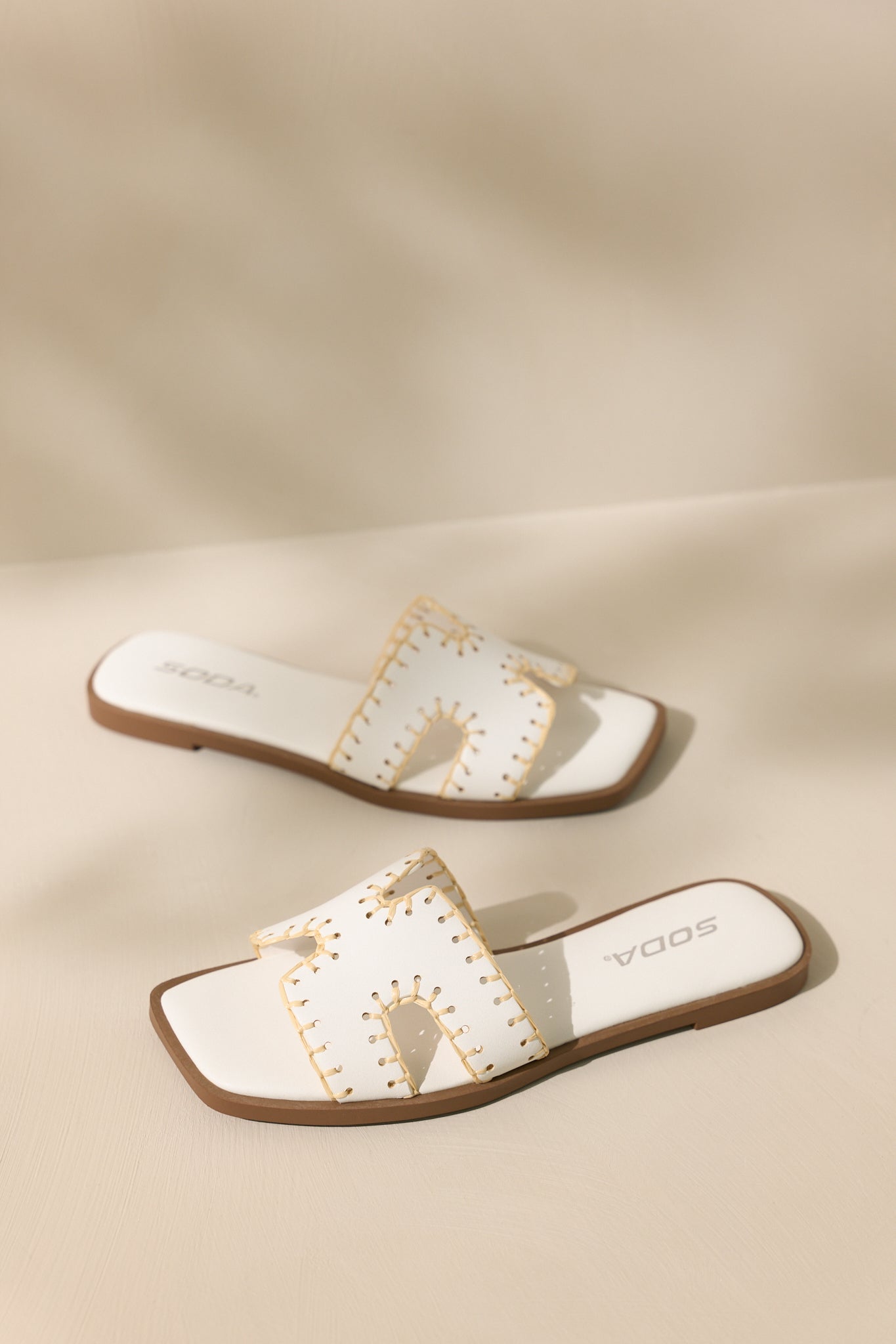 Upper view of these white sandals that feature a square toe, a slip on design, a strap with cutouts over the top of the foot beige stitch detailing, and a high contrasting sole.