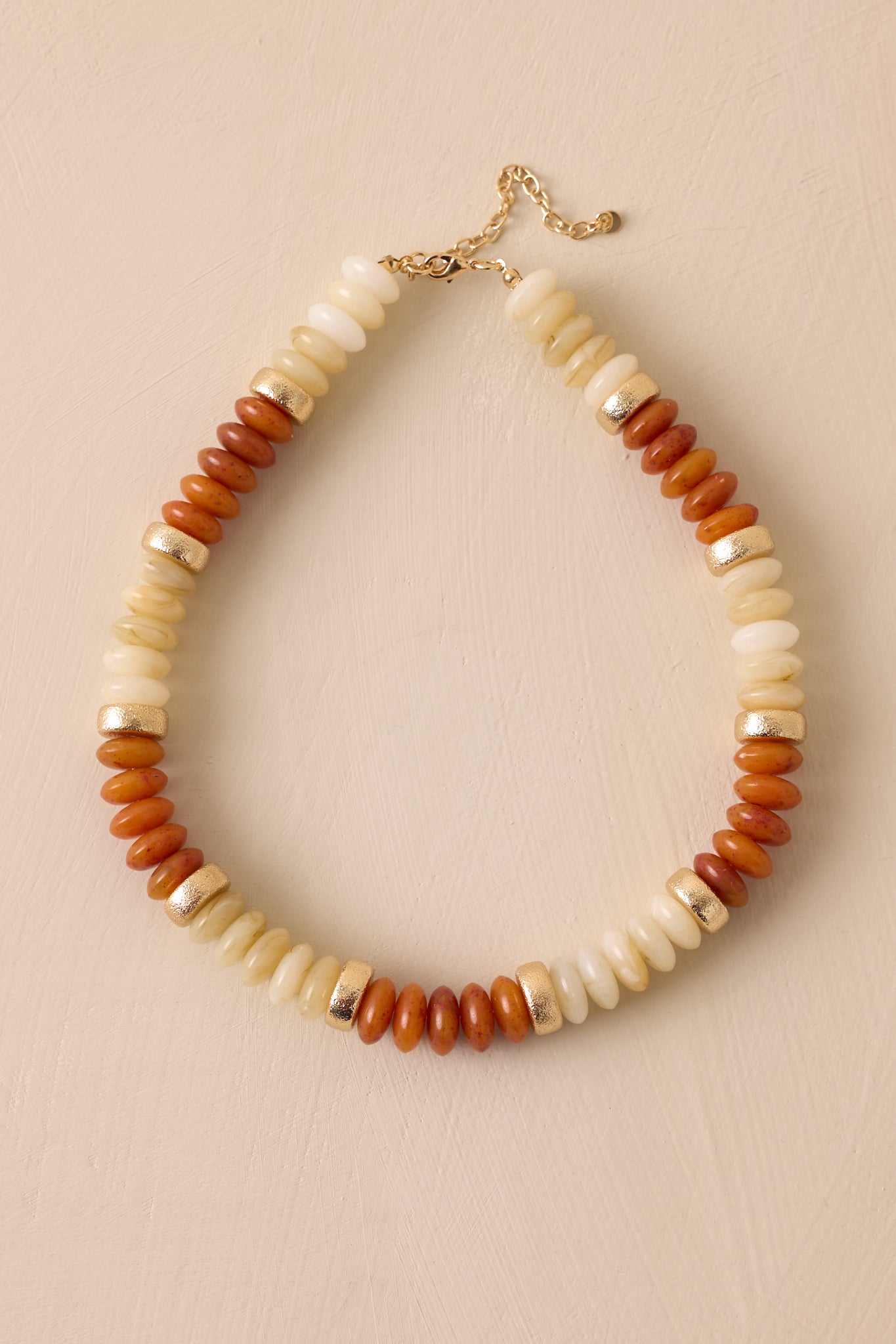 Overhead view of this brown necklace that features gold hardware, ivory, gold, and brown flat beads, and a lobster clasp closure.