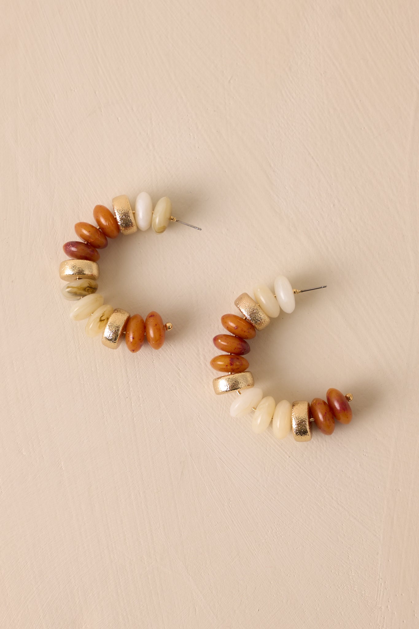 Over-head view of these beaded earrings that feature gold hardware, ivory and brown flat beads with accent gold beads, an incomplete hoop and secure back postings.