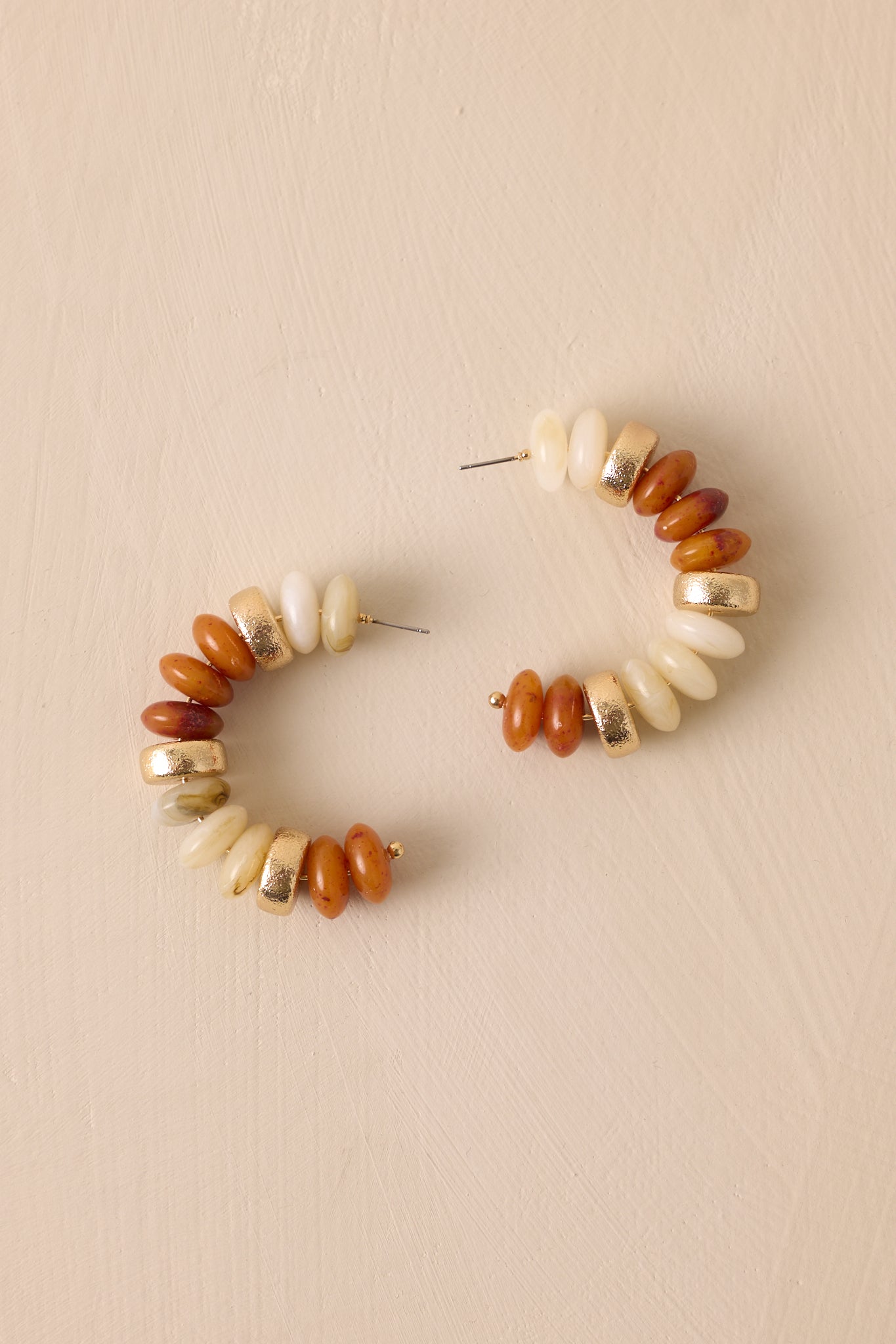 Close-up overhead view of these beaded earrings that feature gold hardware, ivory and brown flat beads with accent gold beads, an incomplete hoop and secure back postings.