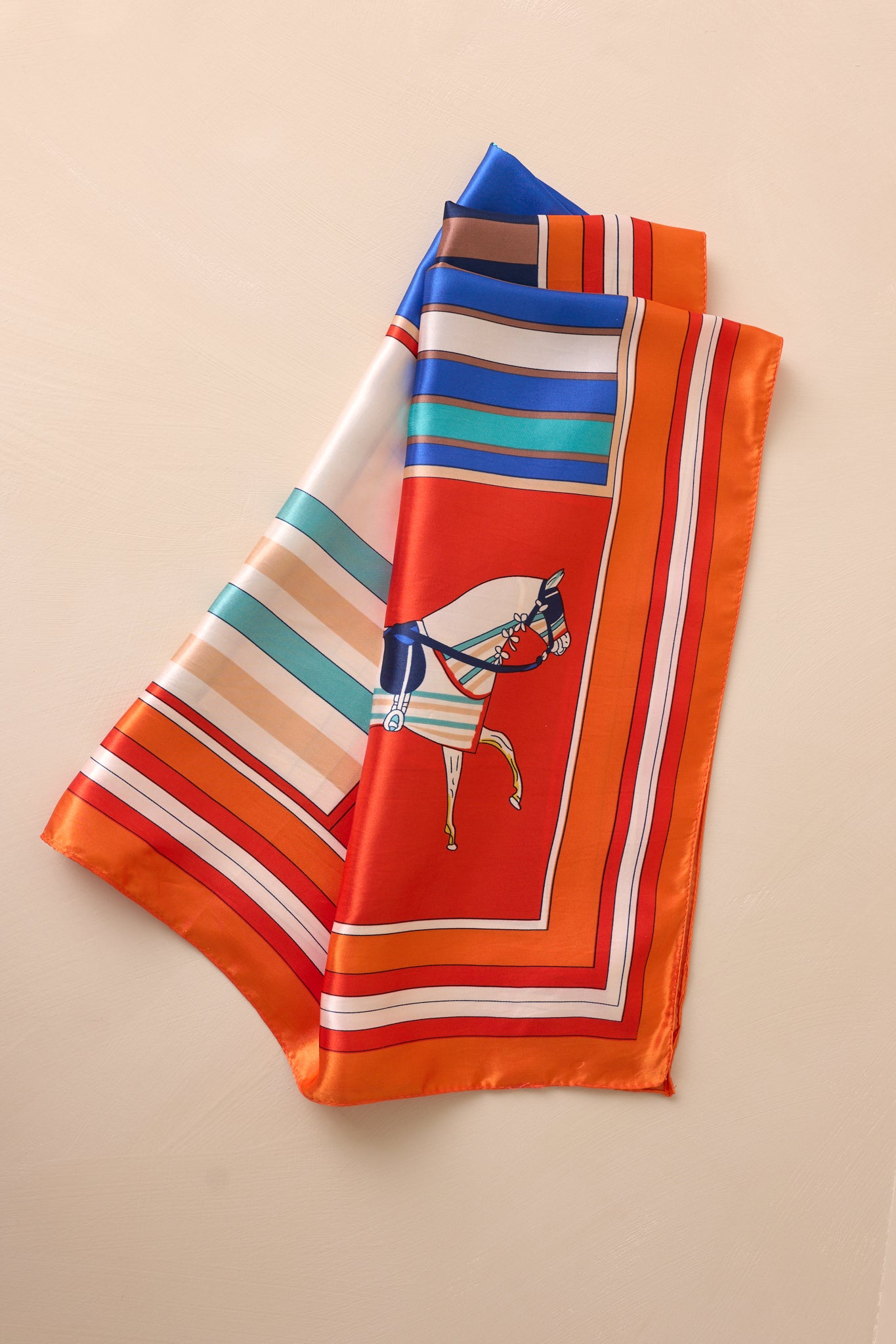 Overhead view of this orange scarf that features an orange trim, with accent colors of ivory, teal, orange and yellow, and a design with horses on it.