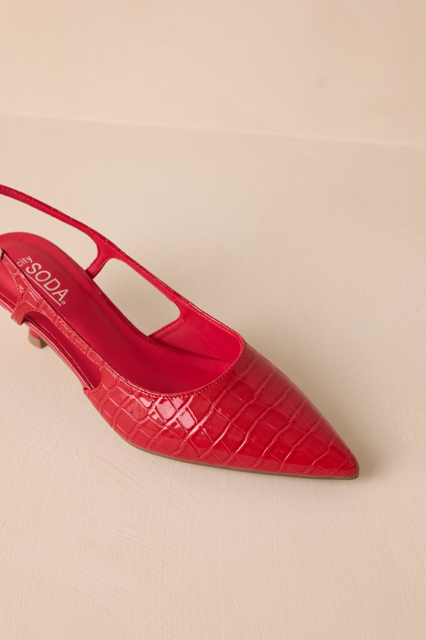 Close-up view of these red heels that feature a small gold buckle detailing, a pointed heel and small cutout detail on the side.