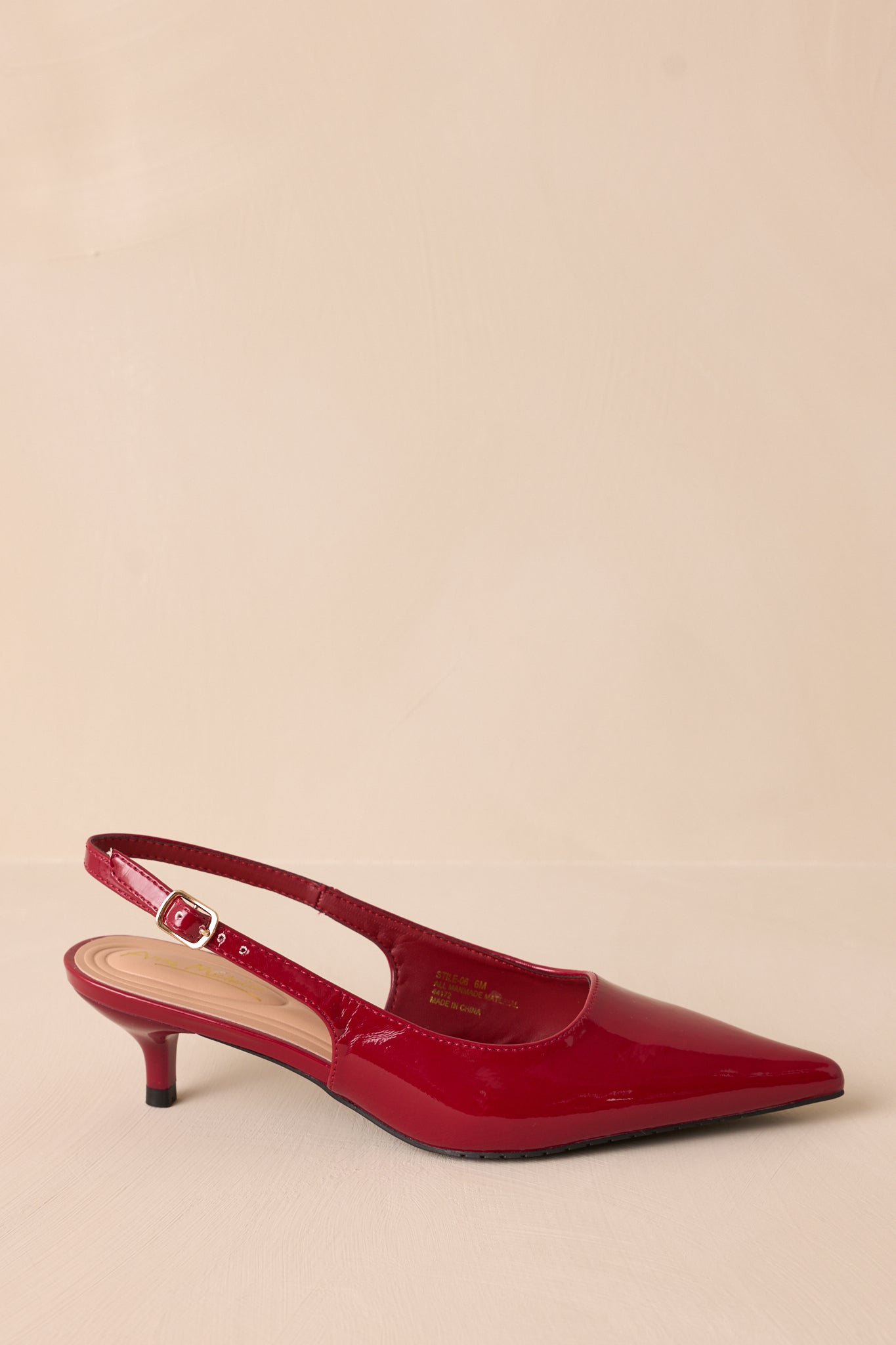 A single burgundy patent slingback heel displayed on a neutral background. The pointed toe, adjustable strap with gold buckle, and short kitten heel are highlighted in this view.