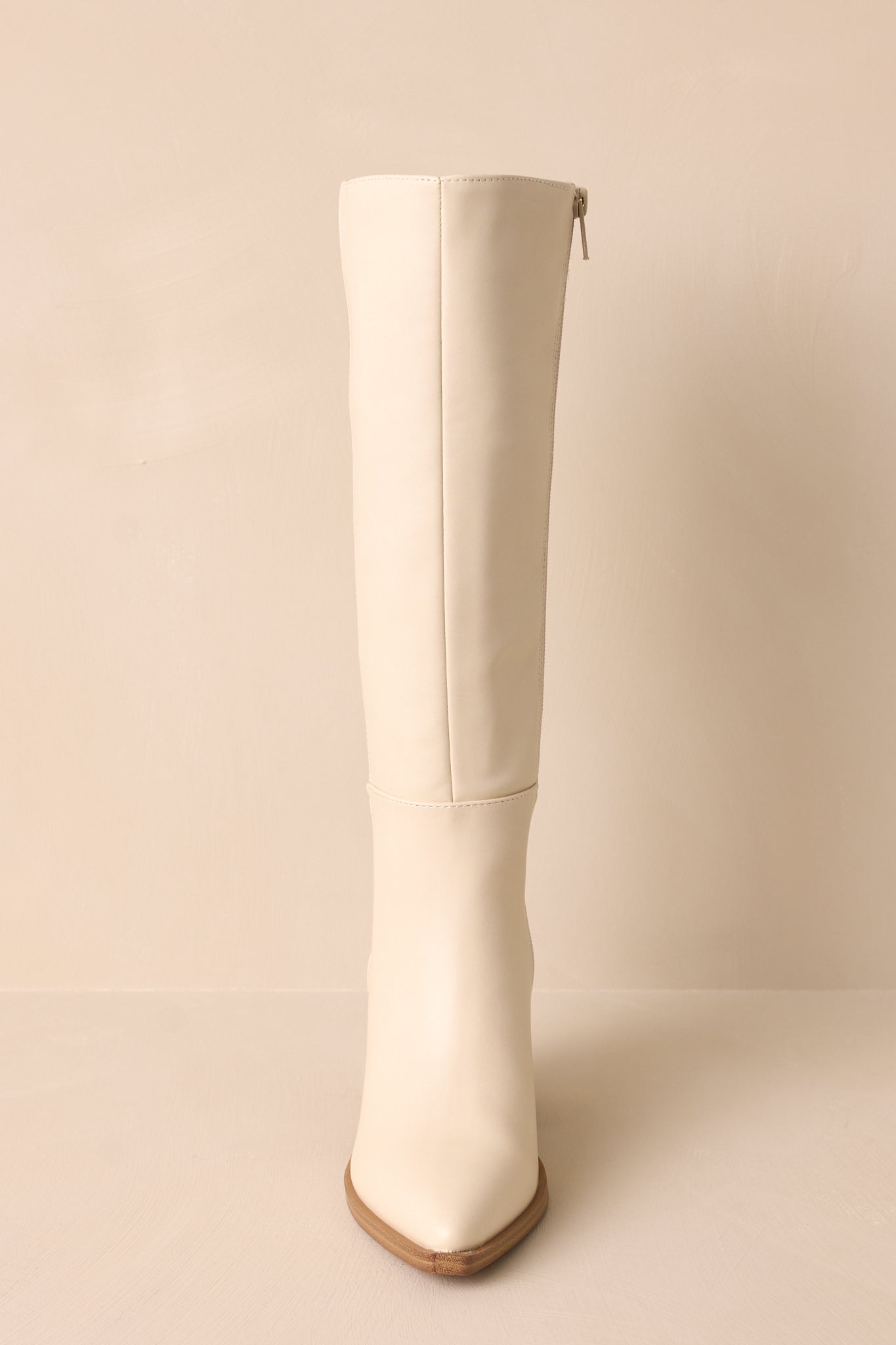 Front view of ivory boots featuring a pointed toe, a sleek faux leather ivory material, a brown block heel, a functional side zipper, a knee high design and a brown sole.