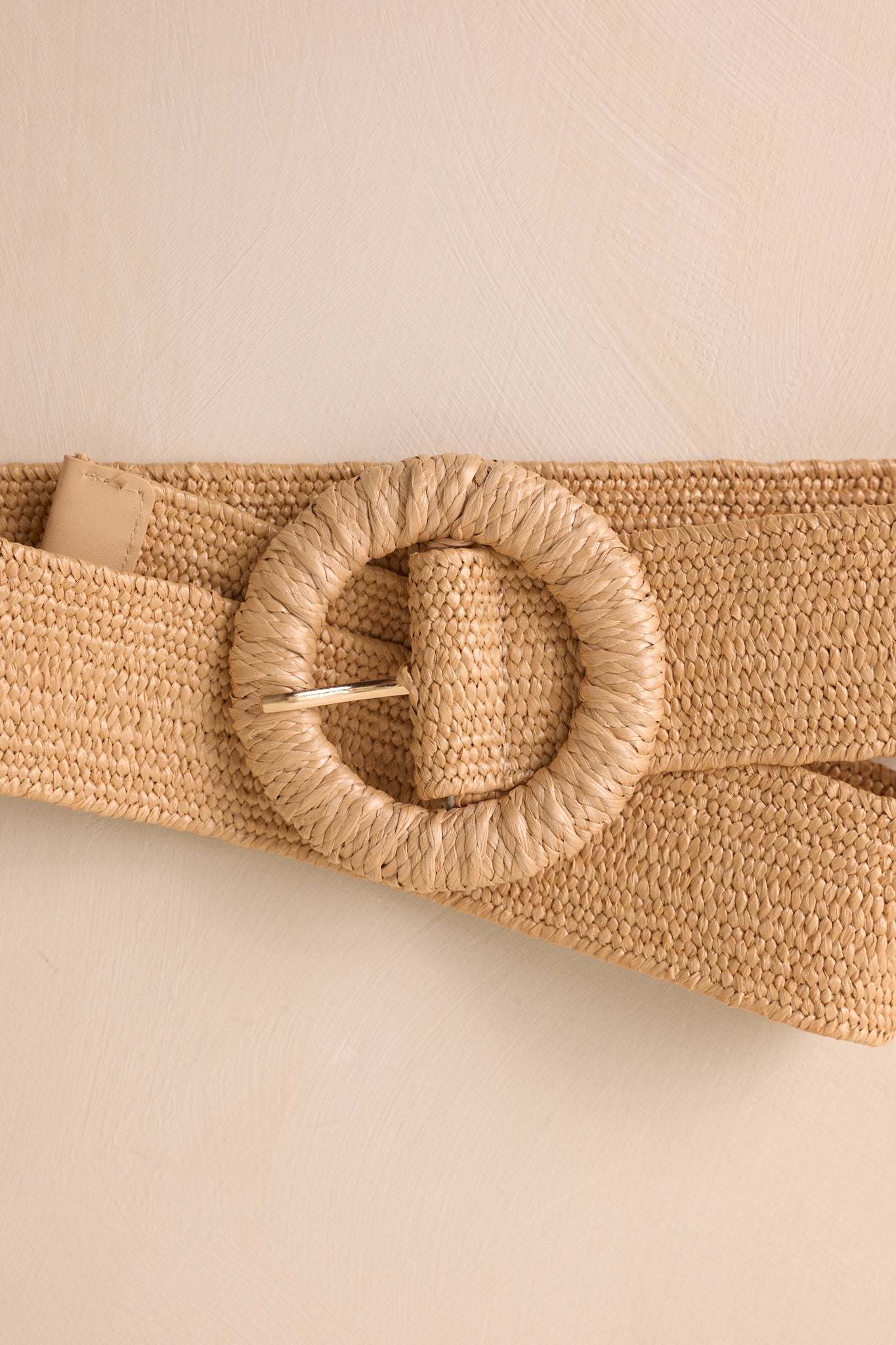 A partial view of the tan belt, showcasing the woven straw design and the round woven belt buckle with gold hardware.