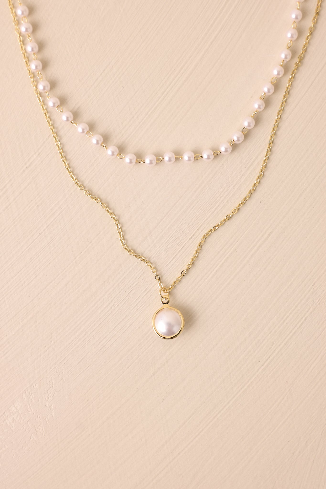 Gold layered necklace with a lobster claw clasp, a shorter strand of faux pearls clasped together, and a second longer strand with a larger faux pearl dangling from the gold chain. 