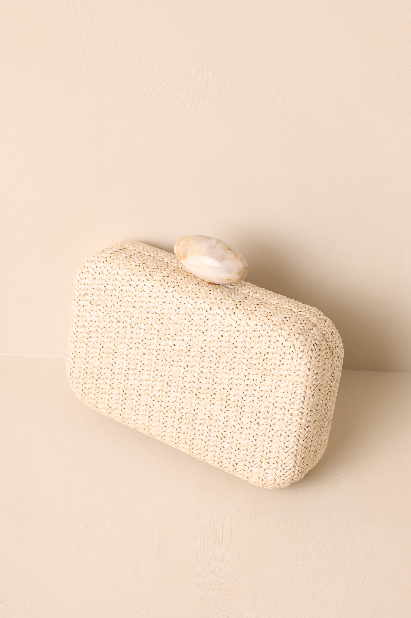 Another angle of the clutch resting on a beige background, highlighting its simple, elegant shape and luxurious finish.