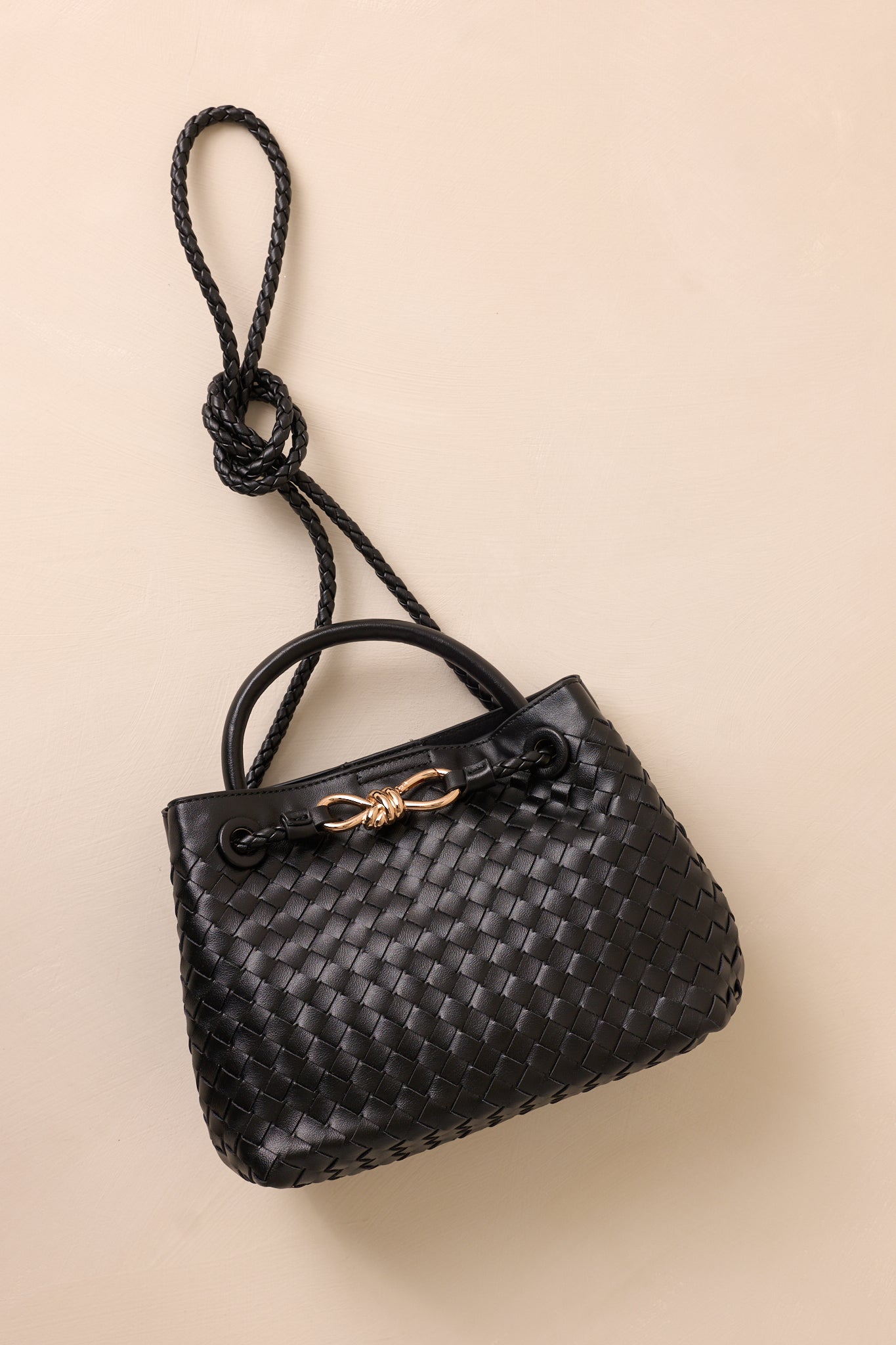 The black faux leather bag laid flat against the beige background, with the long removable woven strap draped across the bag.