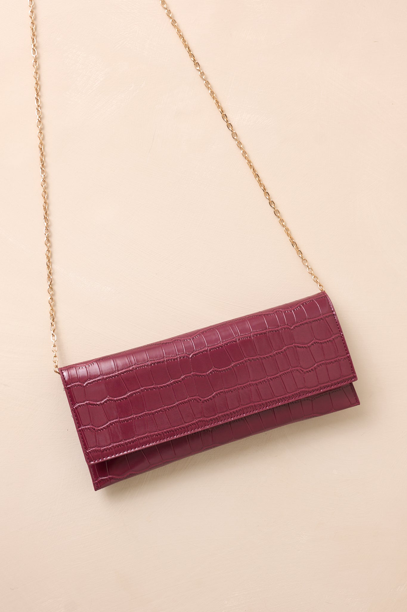 The wine clutch laid flat on a beige surface, with the optional gold chain strap displayed alongside the bag to emphasize its versatility.