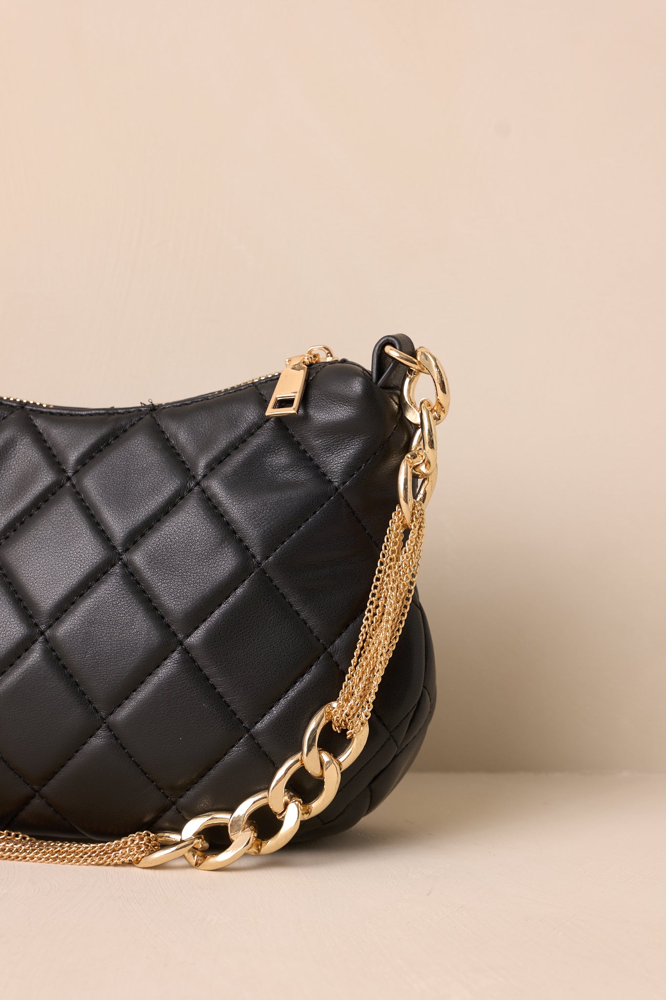 A detailed view of the black faux leather handbag, focusing on the fine quilted stitching, the gleaming gold zipper closure, and the attached thick gold chain handle.