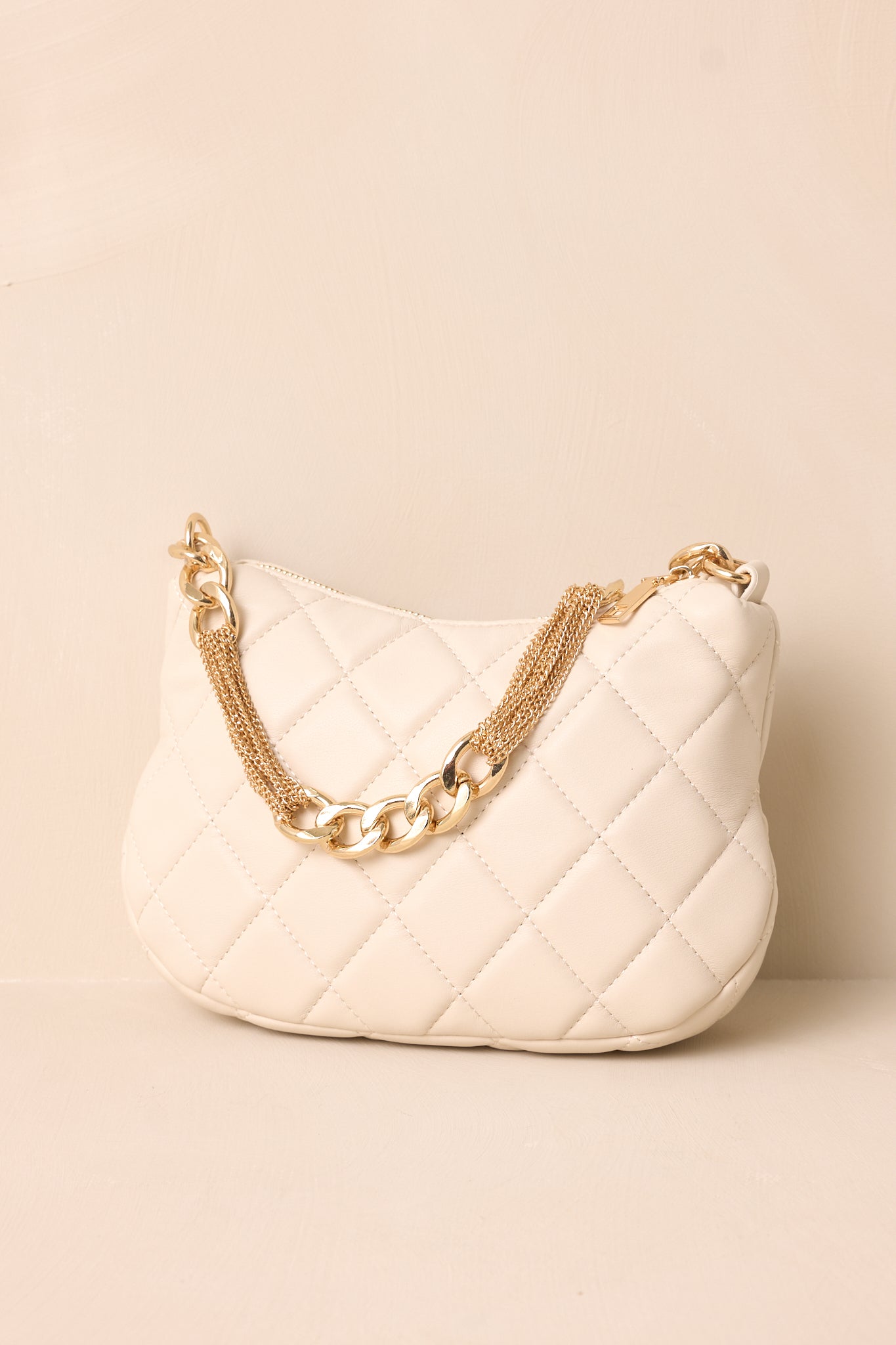 A side angle of the ivory faux leather handbag on a beige background, showing the quilted pattern and the structured silhouette with the thin gold chain shoulder strap resting nearby.