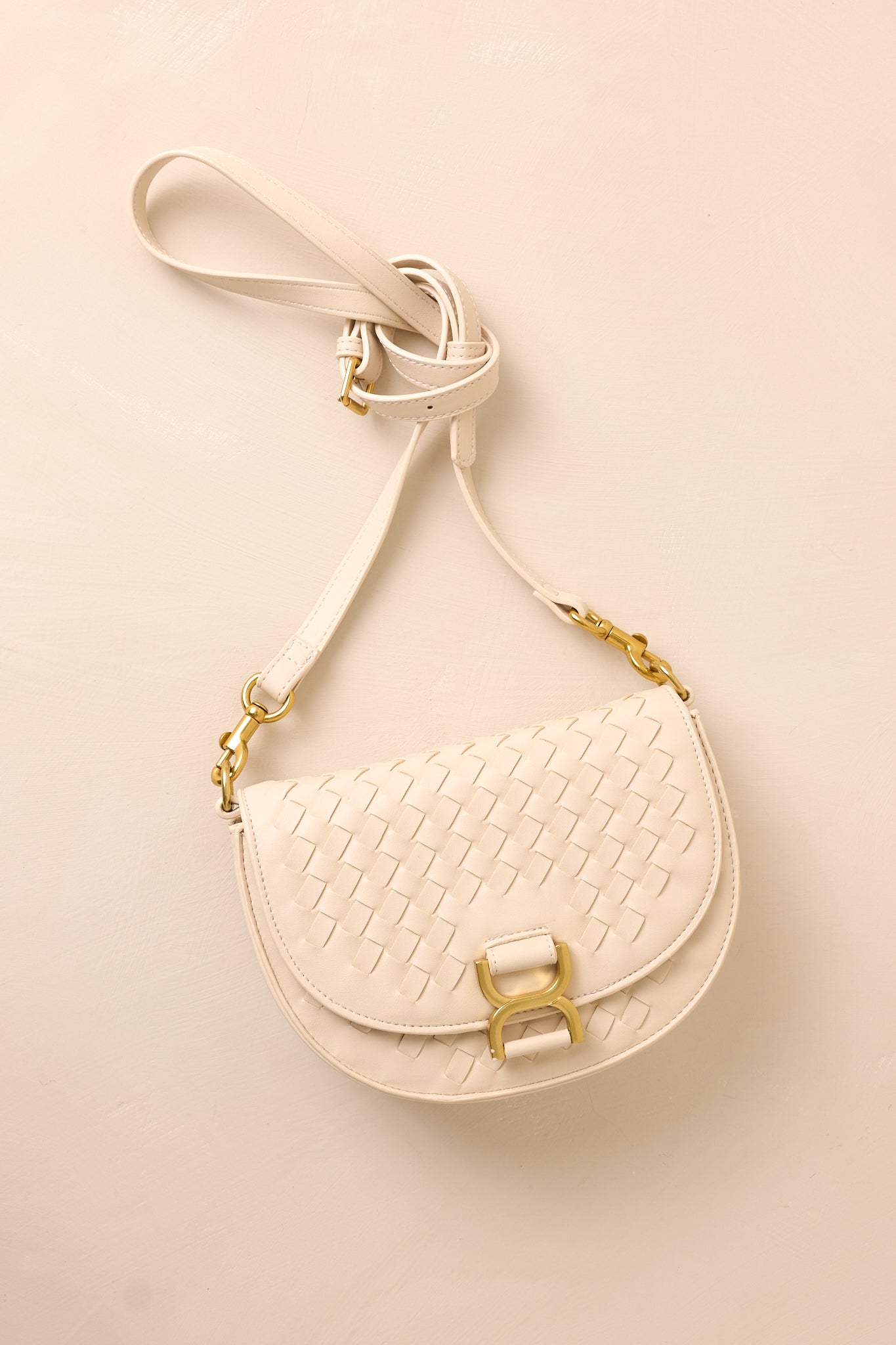 The beige handbag laid flat against a beige background, with the solid beige shoulder strap extended to show its belt-inspired design alongside the gold hardware accents.