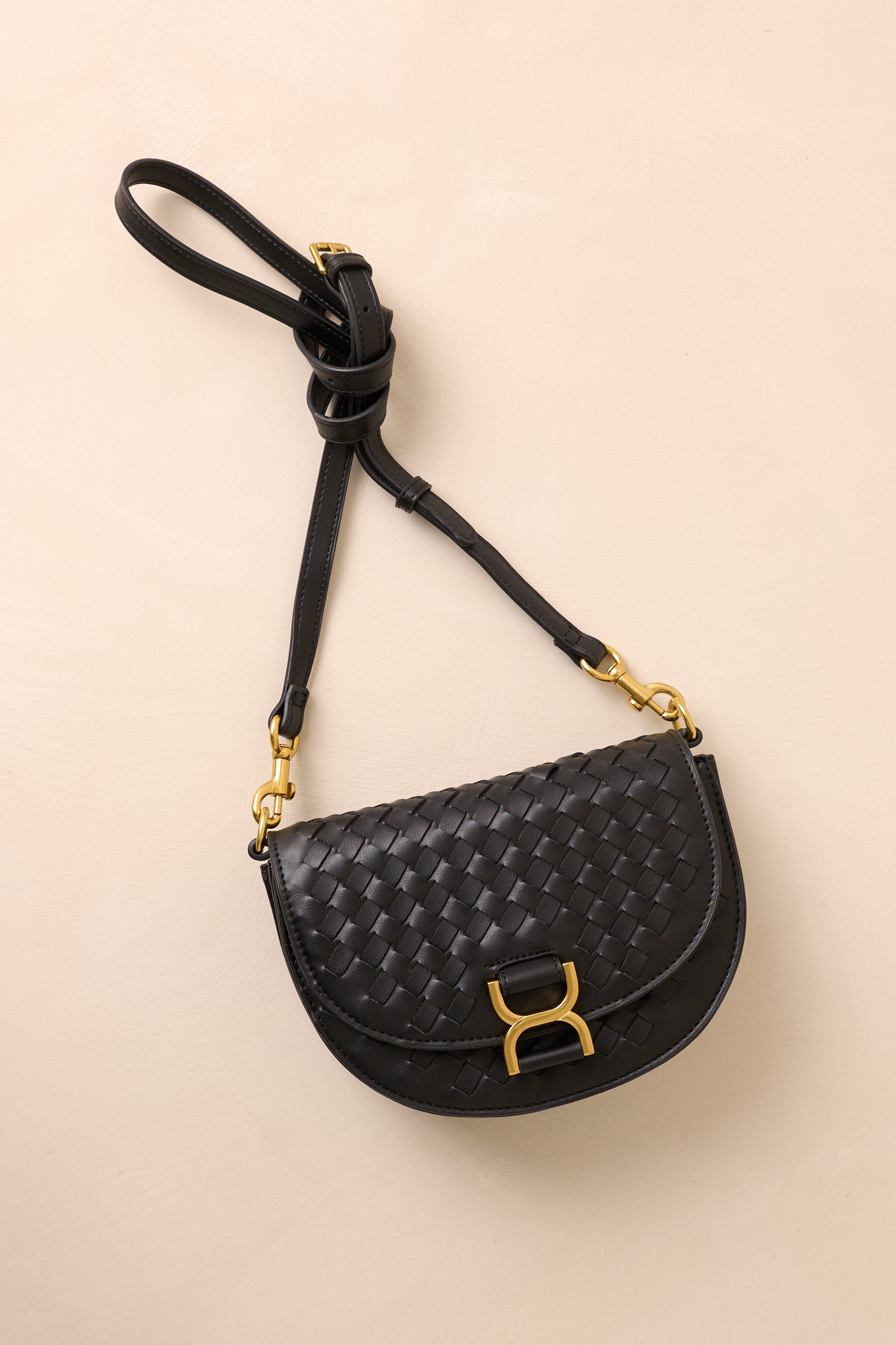 The black handbag laid flat against a beige background, with the solid black shoulder strap extended to show its belt-inspired design alongside the gold hardware accents.