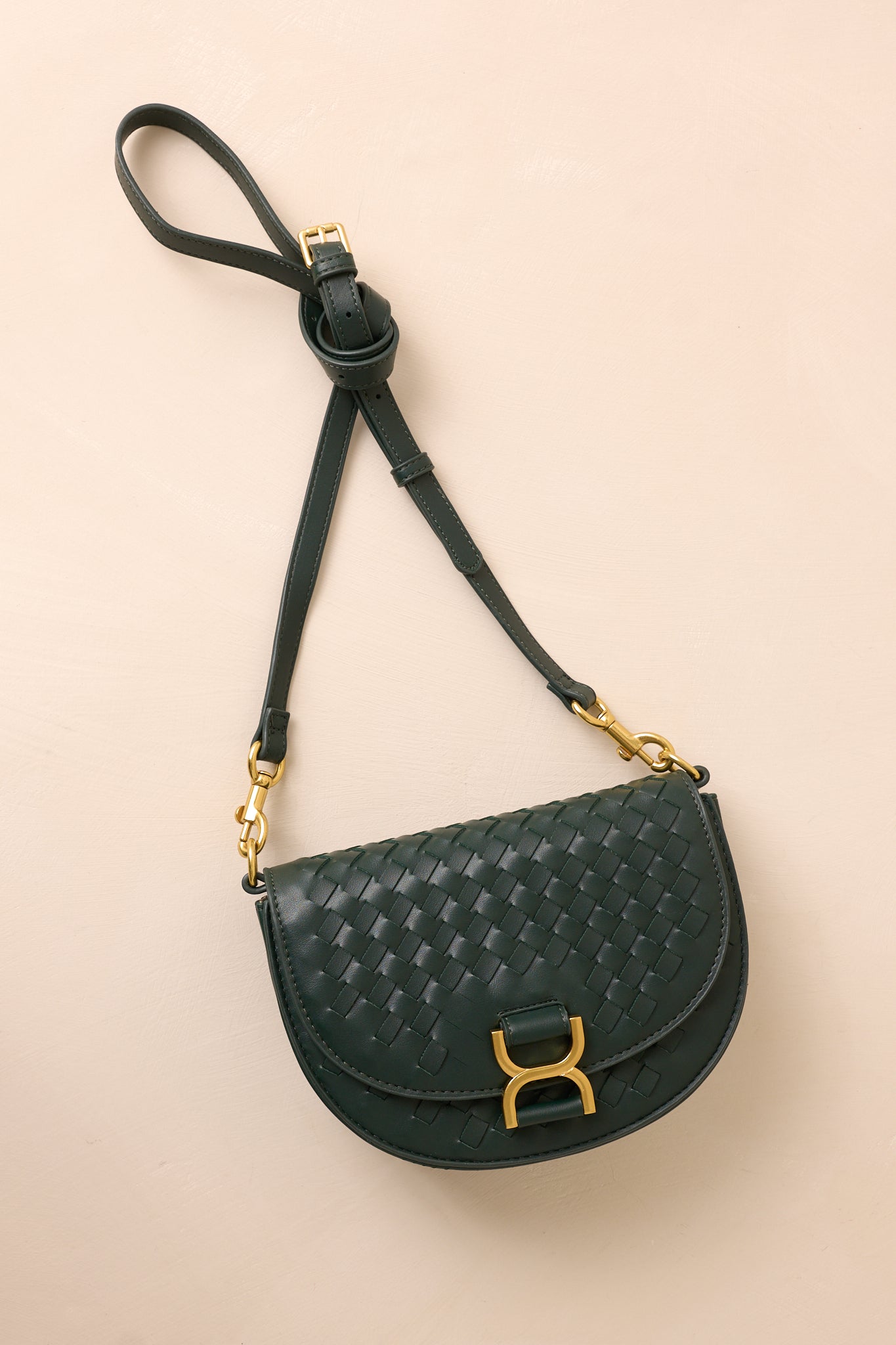 The green handbag laid flat against a beige background, with the solid green shoulder strap extended to show its belt-inspired design alongside the gold hardware accents.