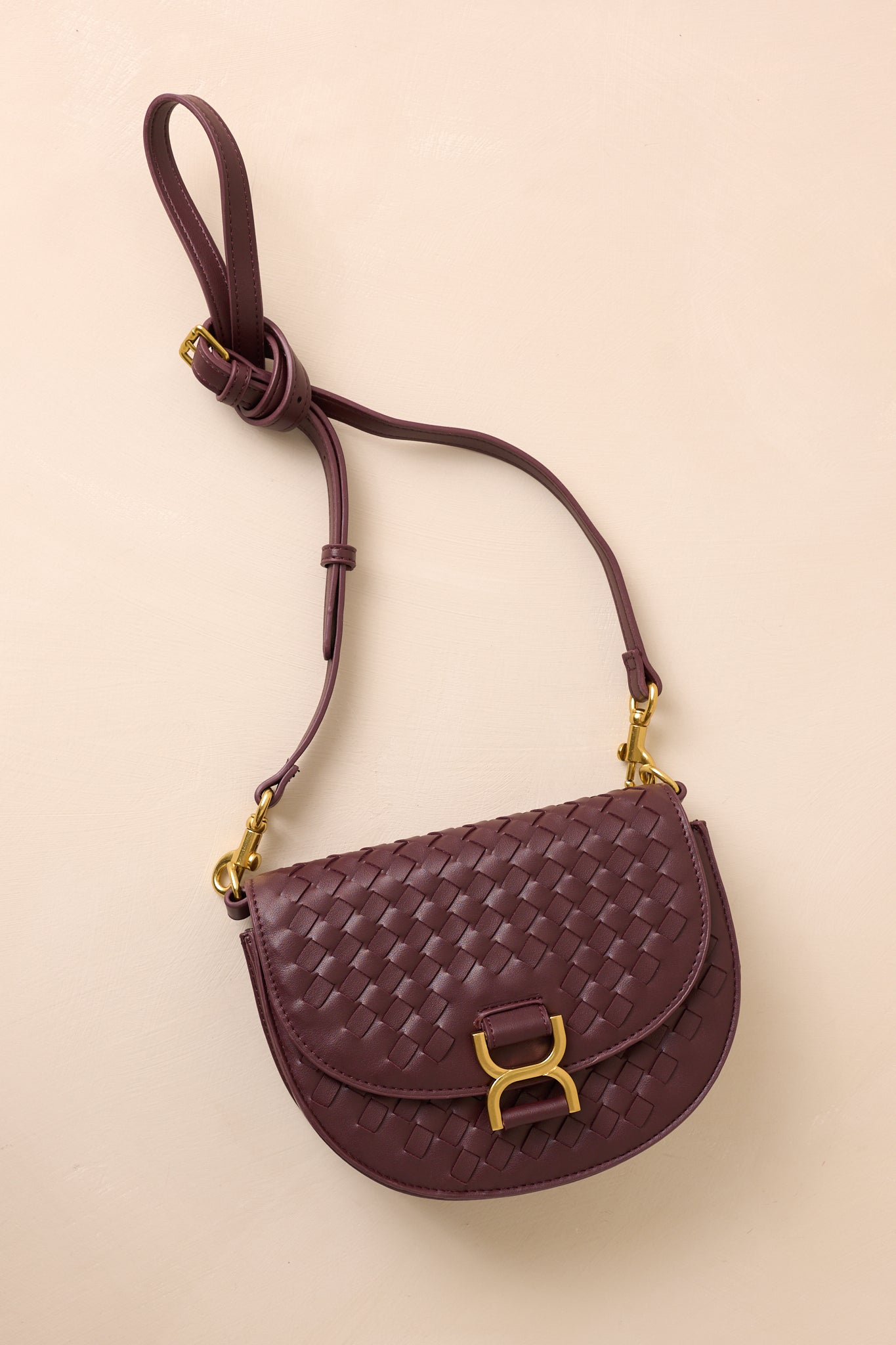 The burgundy handbag laid flat against a beige background, with the solid burgundy shoulder strap extended to show its belt-inspired design alongside the gold hardware accents.