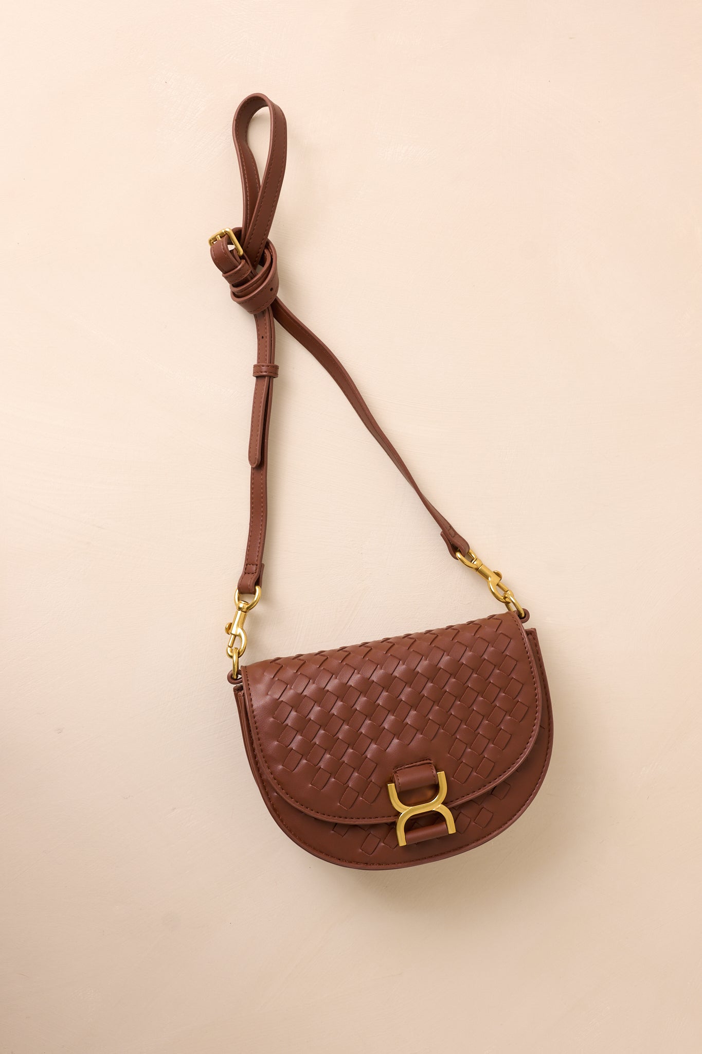 The brown handbag laid flat against a beige background, with the solid brown shoulder strap extended to show its belt-inspired design alongside the gold hardware accents.