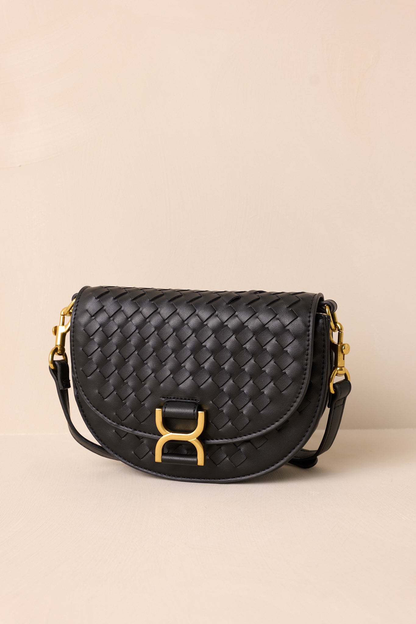 The black handbag displayed upright against a beige background, showcasing its fold-over design, gold buckle snap closure, and woven faux leather material.