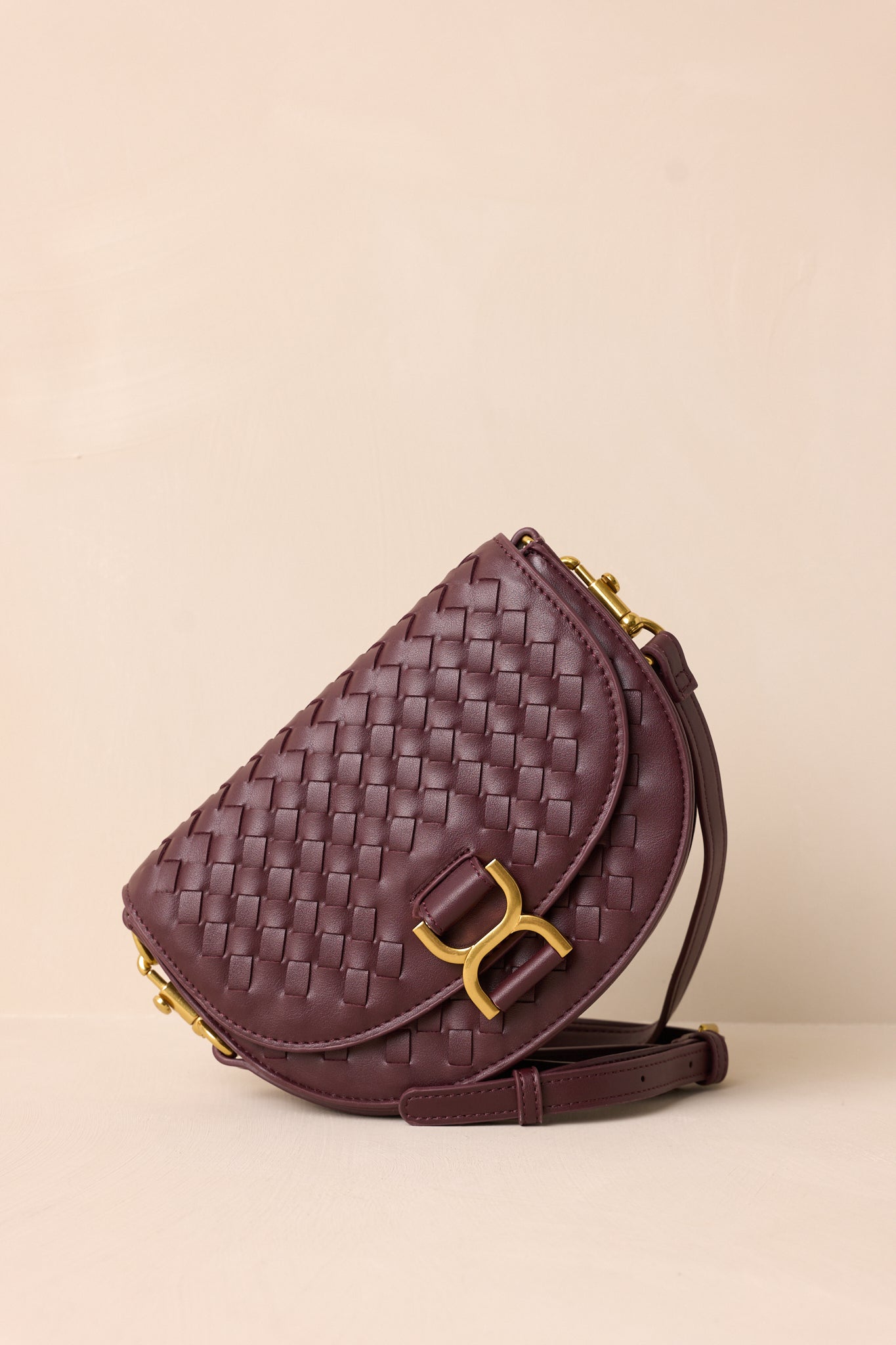 A side angle of the burgundy handbag on a beige background, highlighting the solid burgundy belt-style shoulder strap and the structured silhouette of the design.