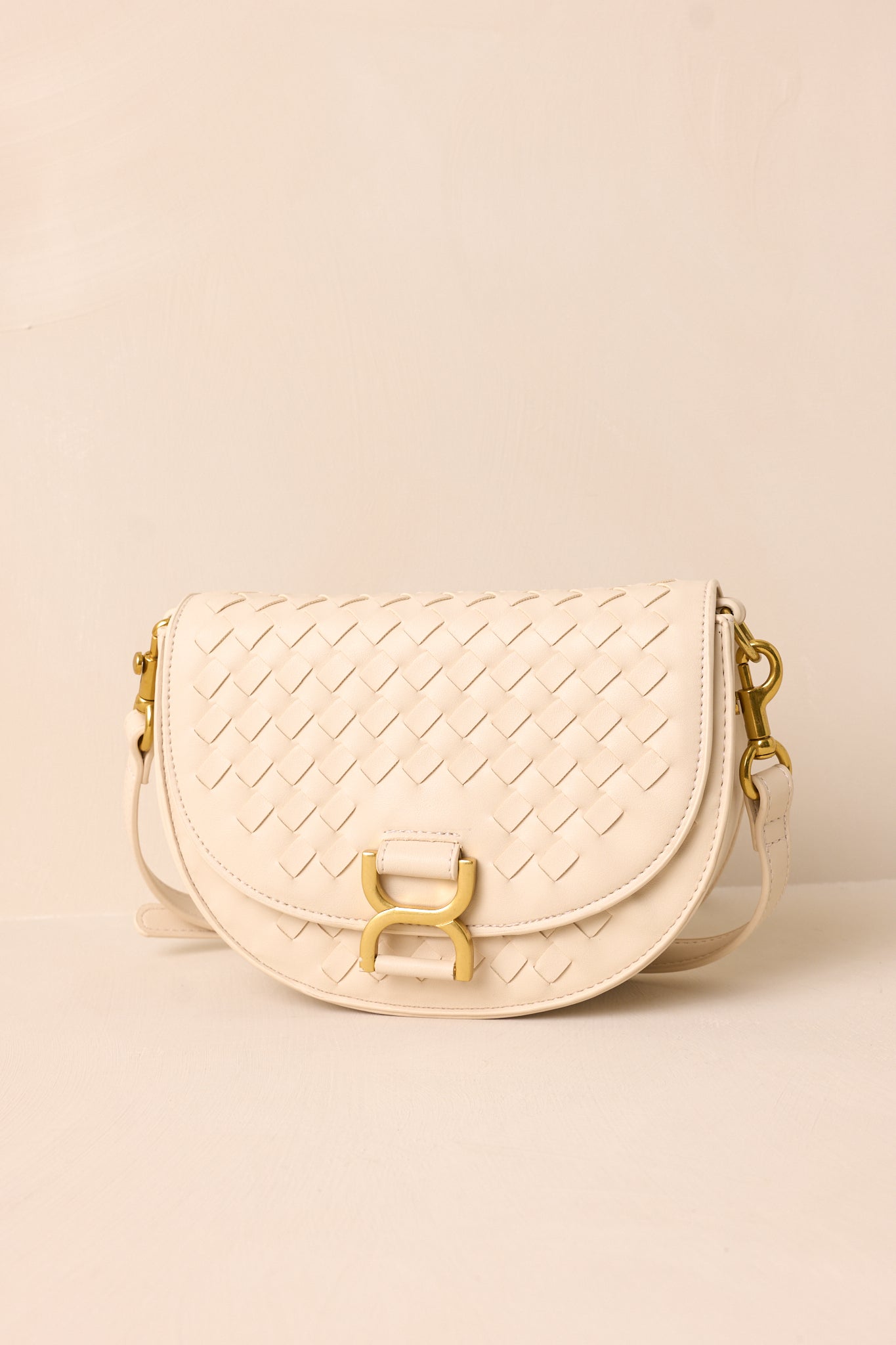 The beige handbag displayed upright against a beige background, showcasing its fold-over design, gold buckle snap closure, and woven faux leather material.