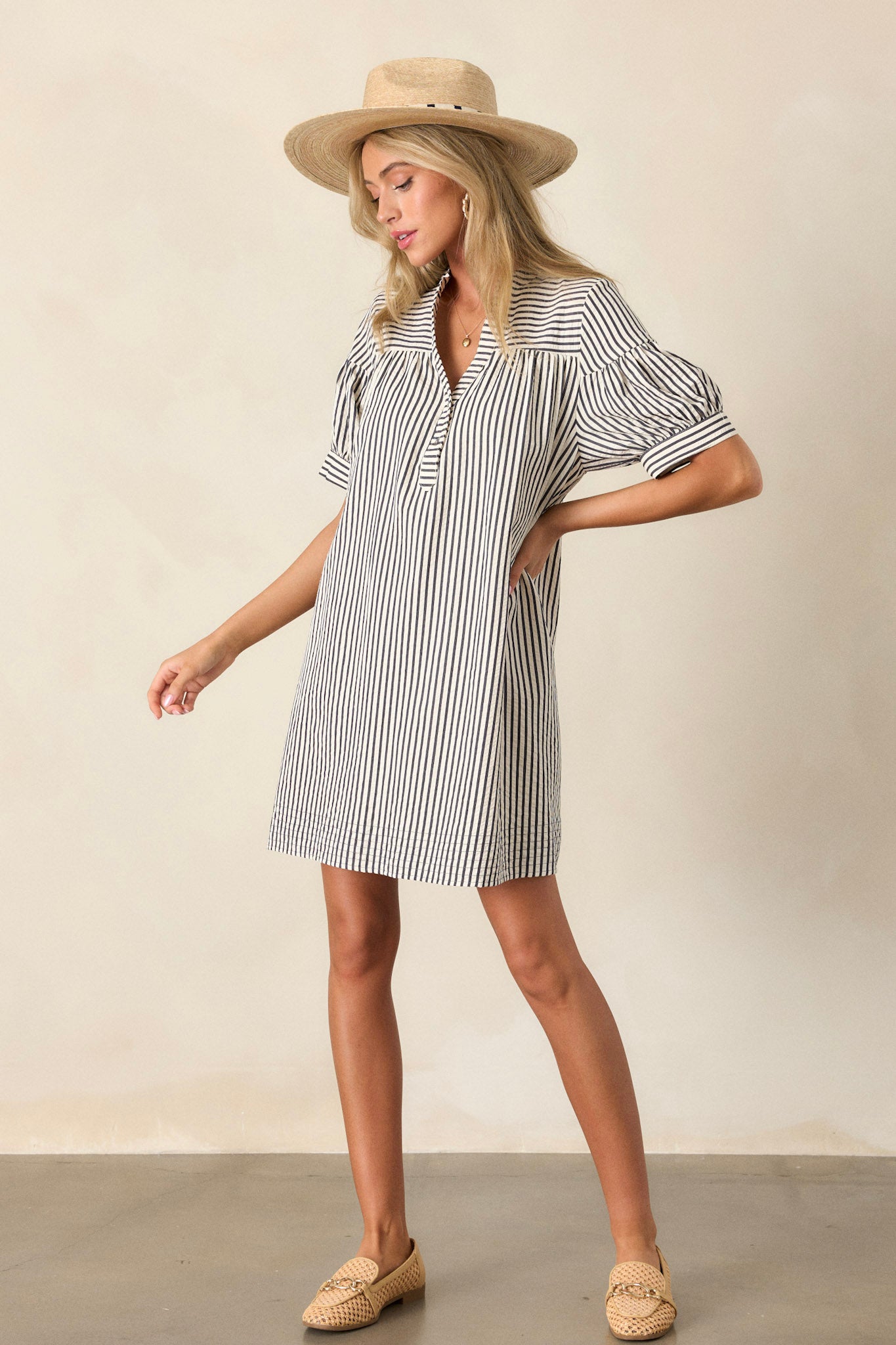 Full body view of this mini dress featuring a v-neckline, a button up bust, a relaxed fit, a vertical navy and white stripe design, and short sleeves.