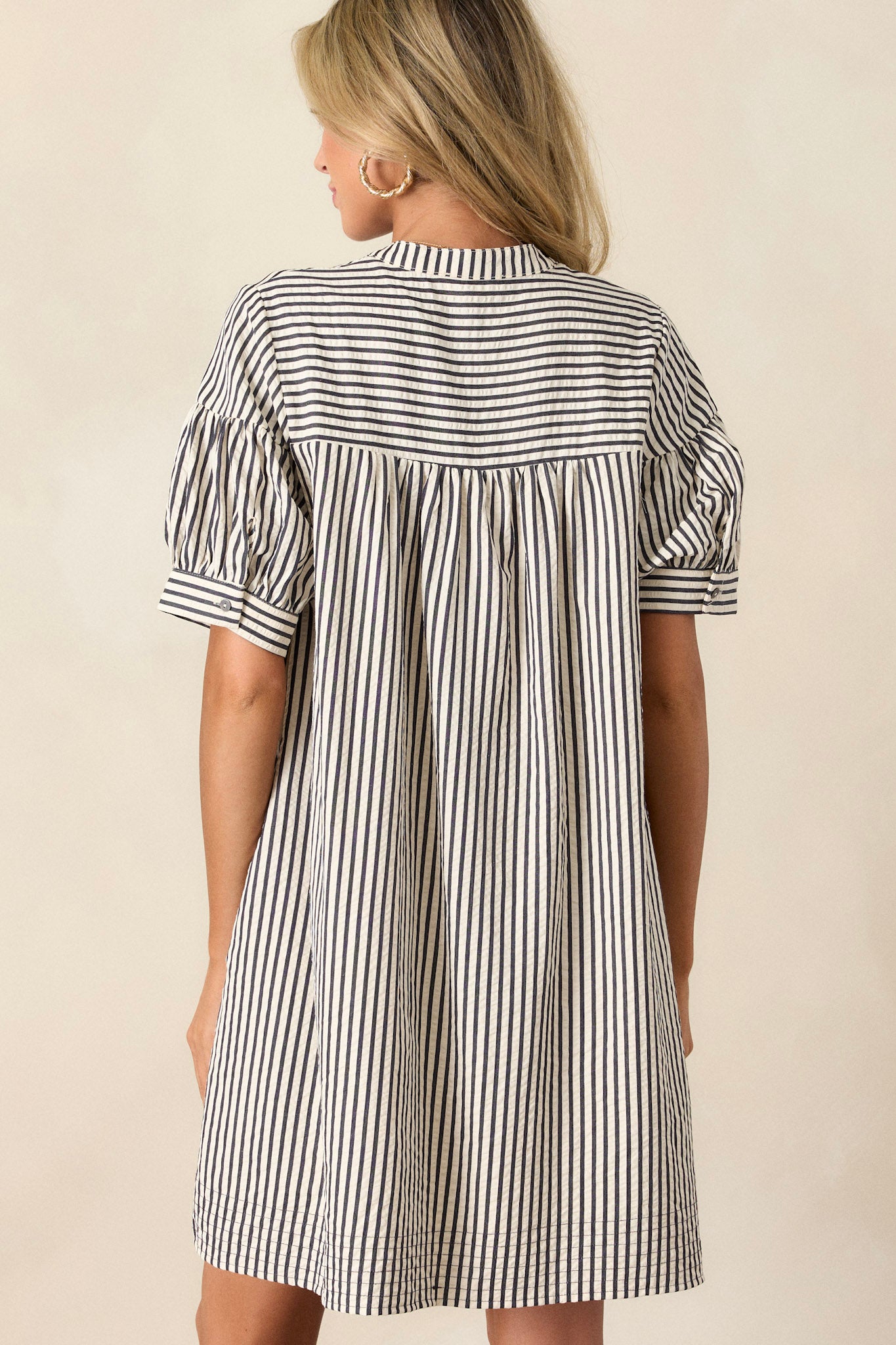 Back view of this mini dress featuring a v-neckline, a button up bust, a relaxed fit, a vertical navy and white stripe design, and short sleeves.