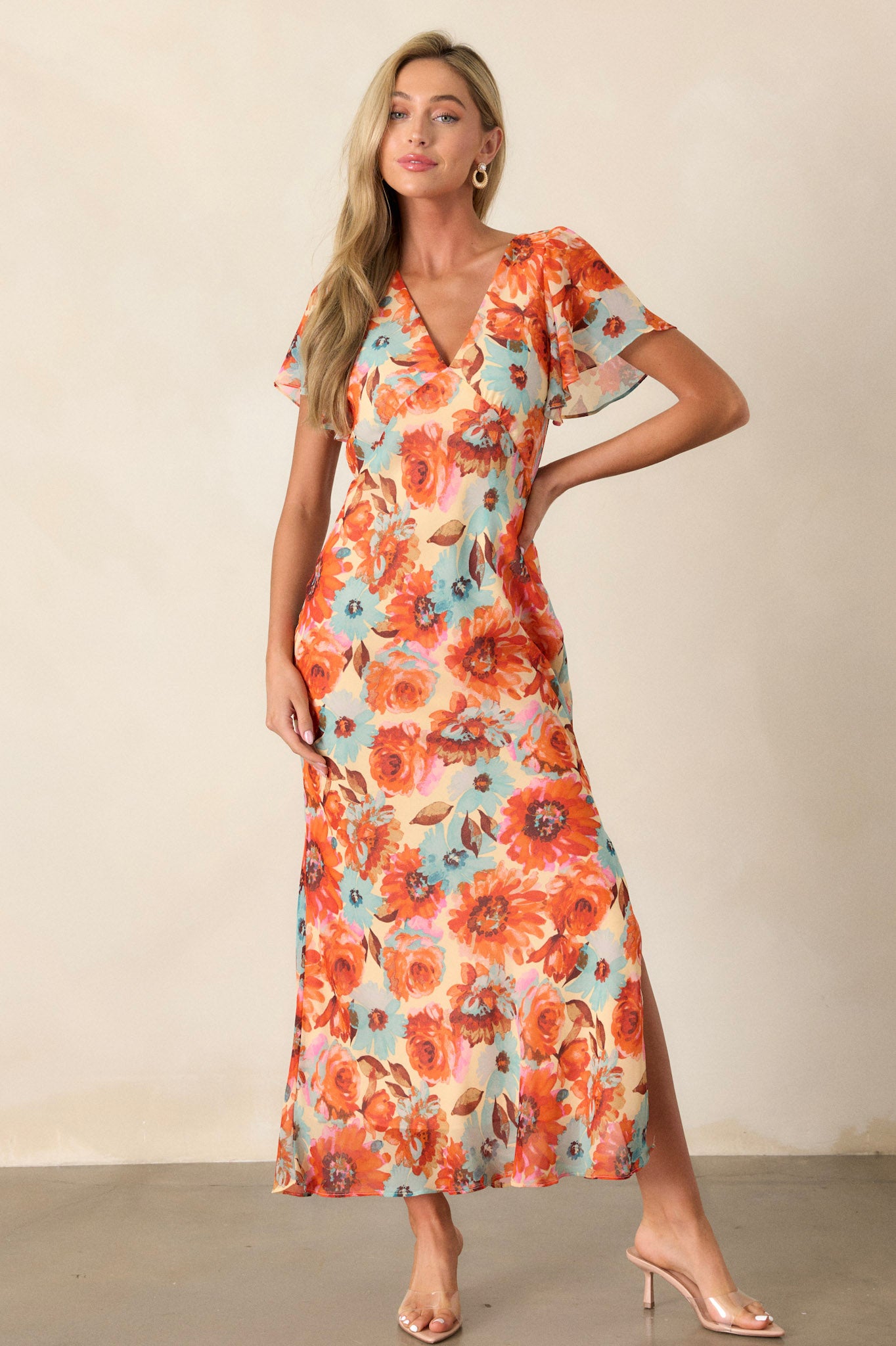 Front view of an orange floral dress featuring a v-neckline, a floral print design, a leg slit on the left leg, an elastic waist insert, a discrete back zipper, and sheer short sleeves.