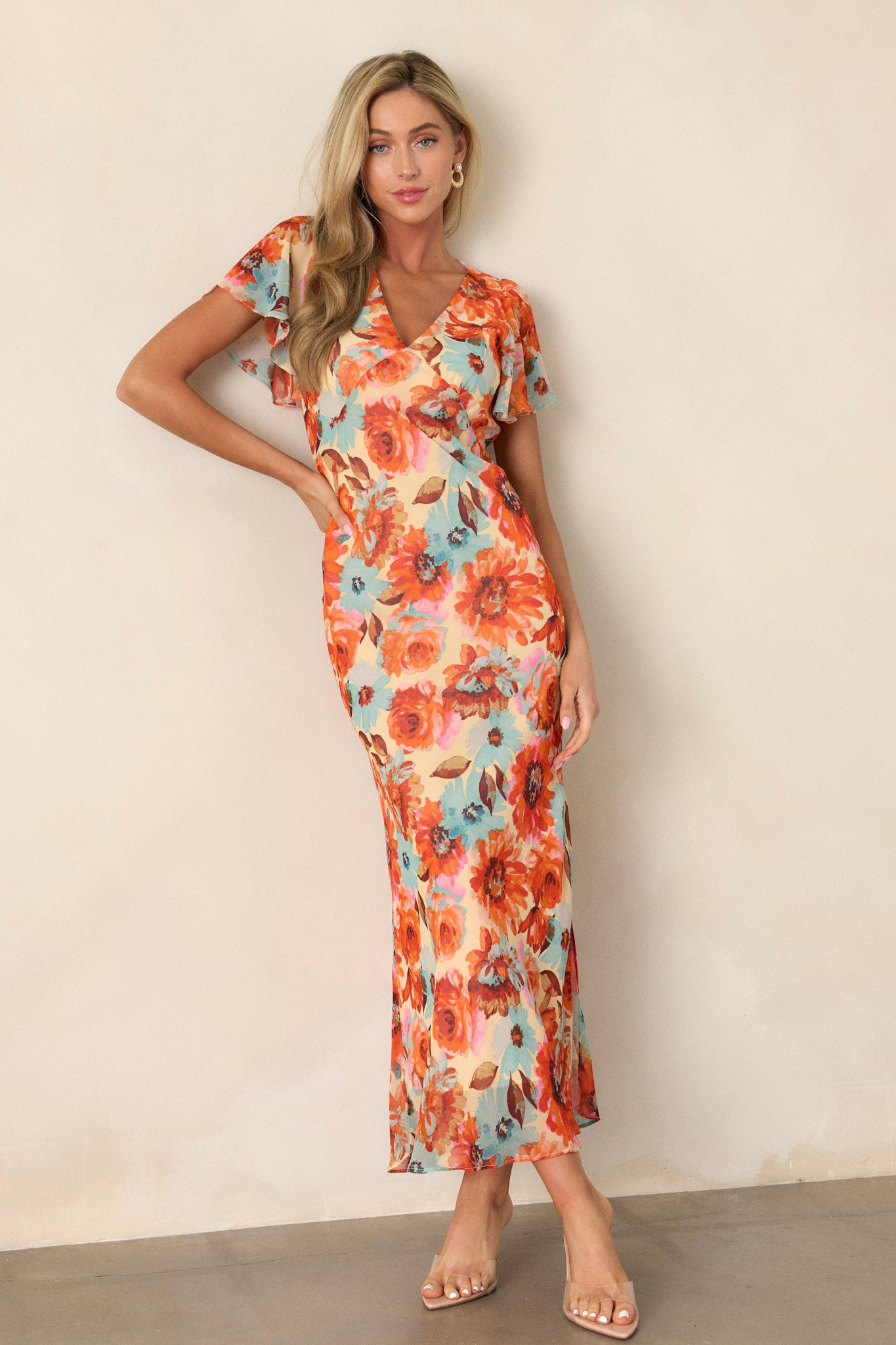 Front angled view of an orange floral dress featuring a v-neckline, floral print design, a leg slit on the left leg, an elastic waist insert, a discrete back zipper, and sheer short sleeves