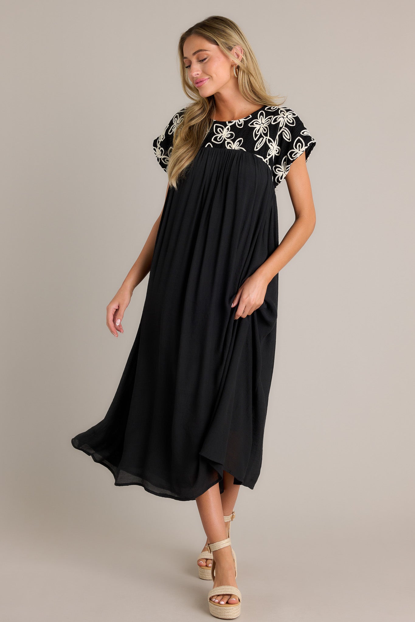 Black midi dress highlighting the high rounded neckline, embroidered floral pattern, and short sleeves with a flowing silhouette.