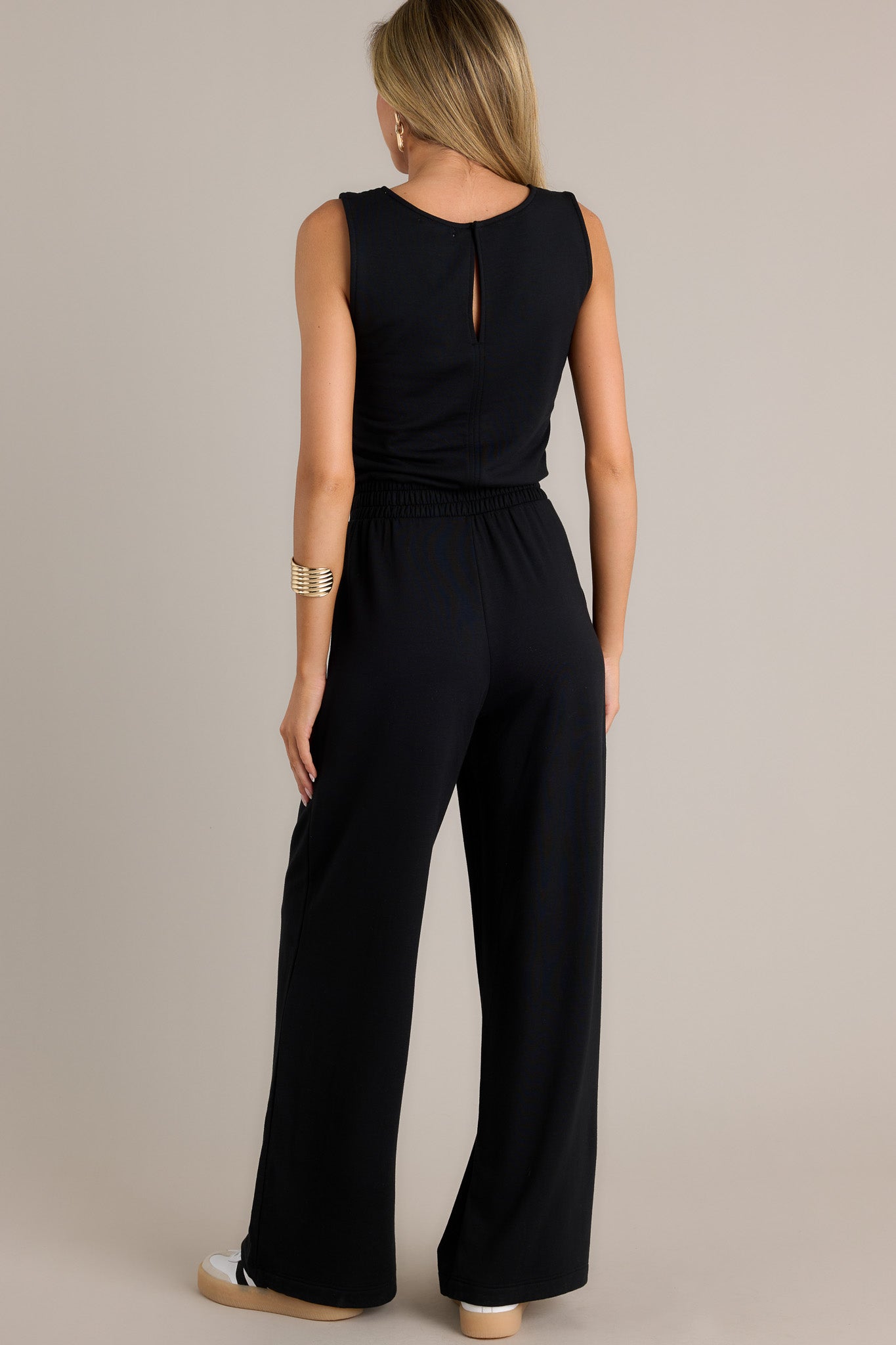 Back view of this black jumpsuit featuring a scoop neckline, a thick elastic waistband, a self-tie drawstring, functional hip pockets, a wide leg, and a sleeveless design.