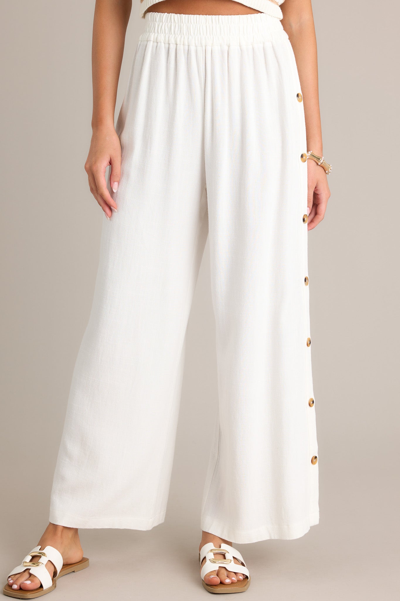 Close up view of these ivory pants featuring a high waisted design, an elastic waistband, functional buttons all down the sides, a lightweight material, and a wide leg.