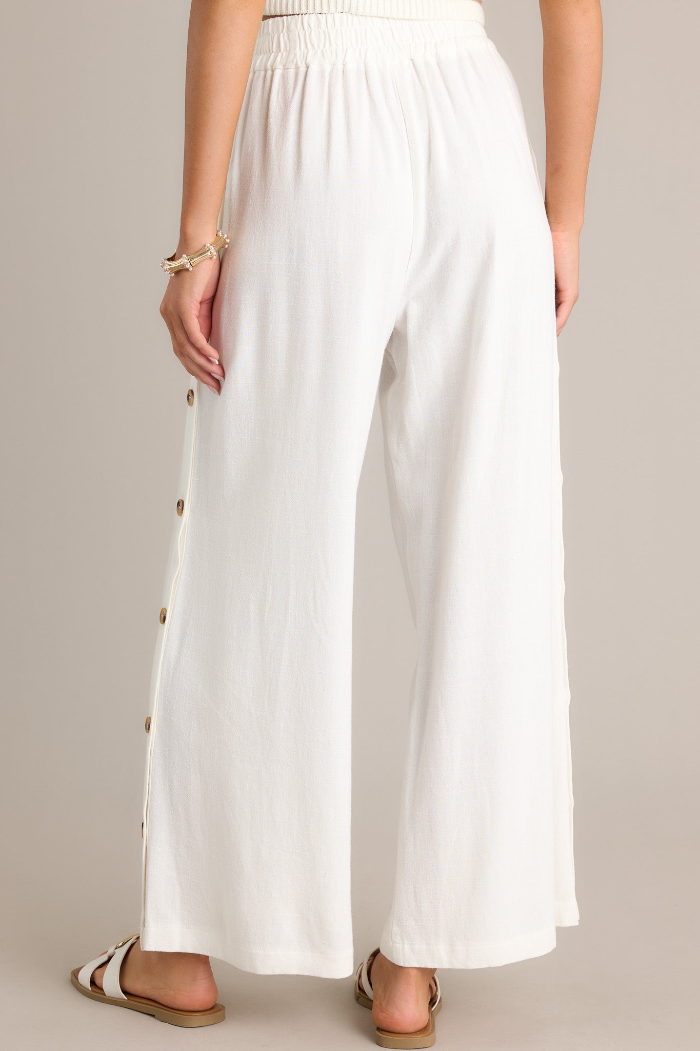 Back view of these ivory pants featuring a high waisted design, an elastic waistband, functional buttons all down the sides, a lightweight material, and a wide leg.