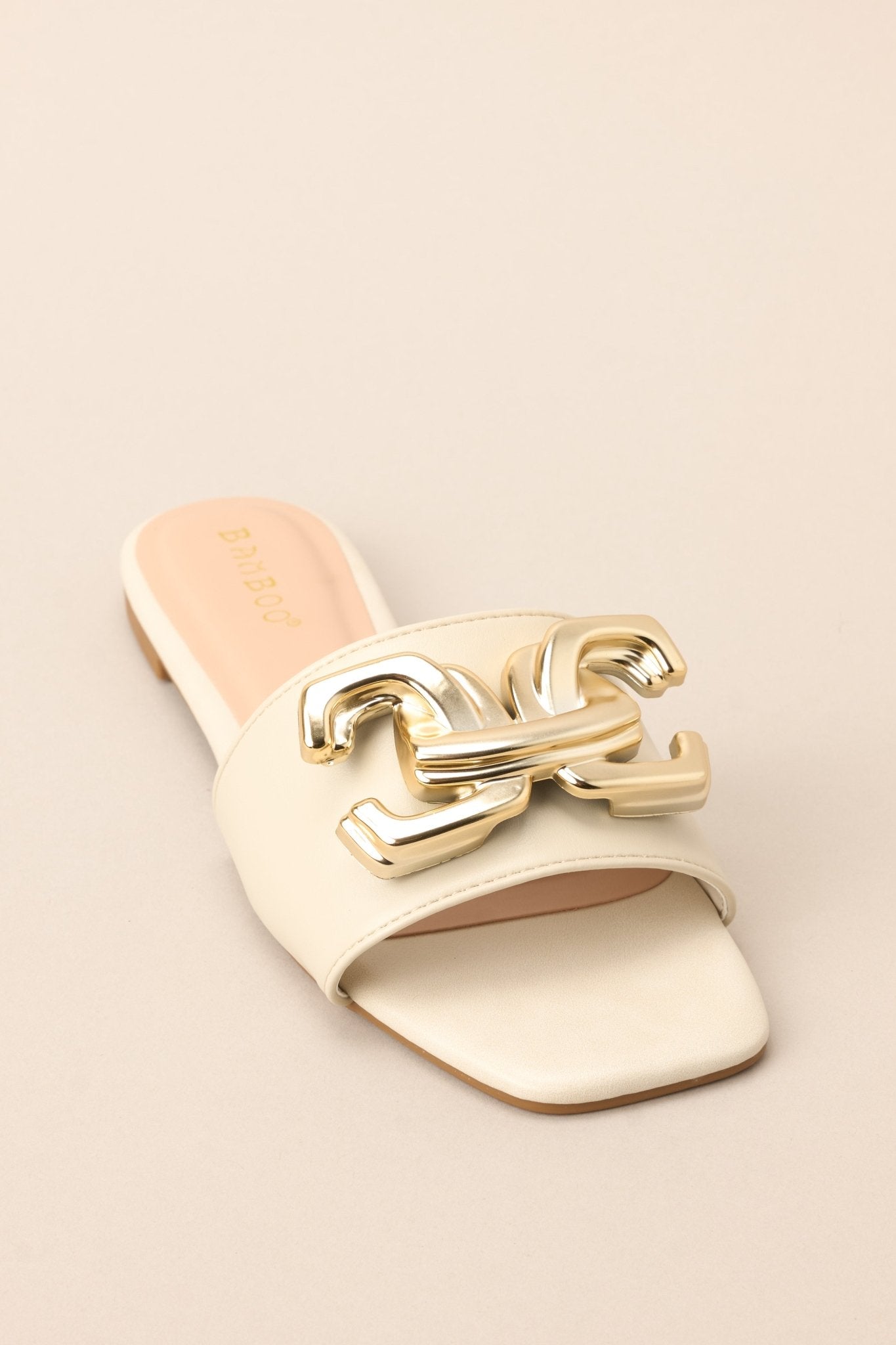 Front angled view of these ivory slip-on sandals that are adorned with a striking large gold buckle-like design, adding a touch of glamour to your ensemble.