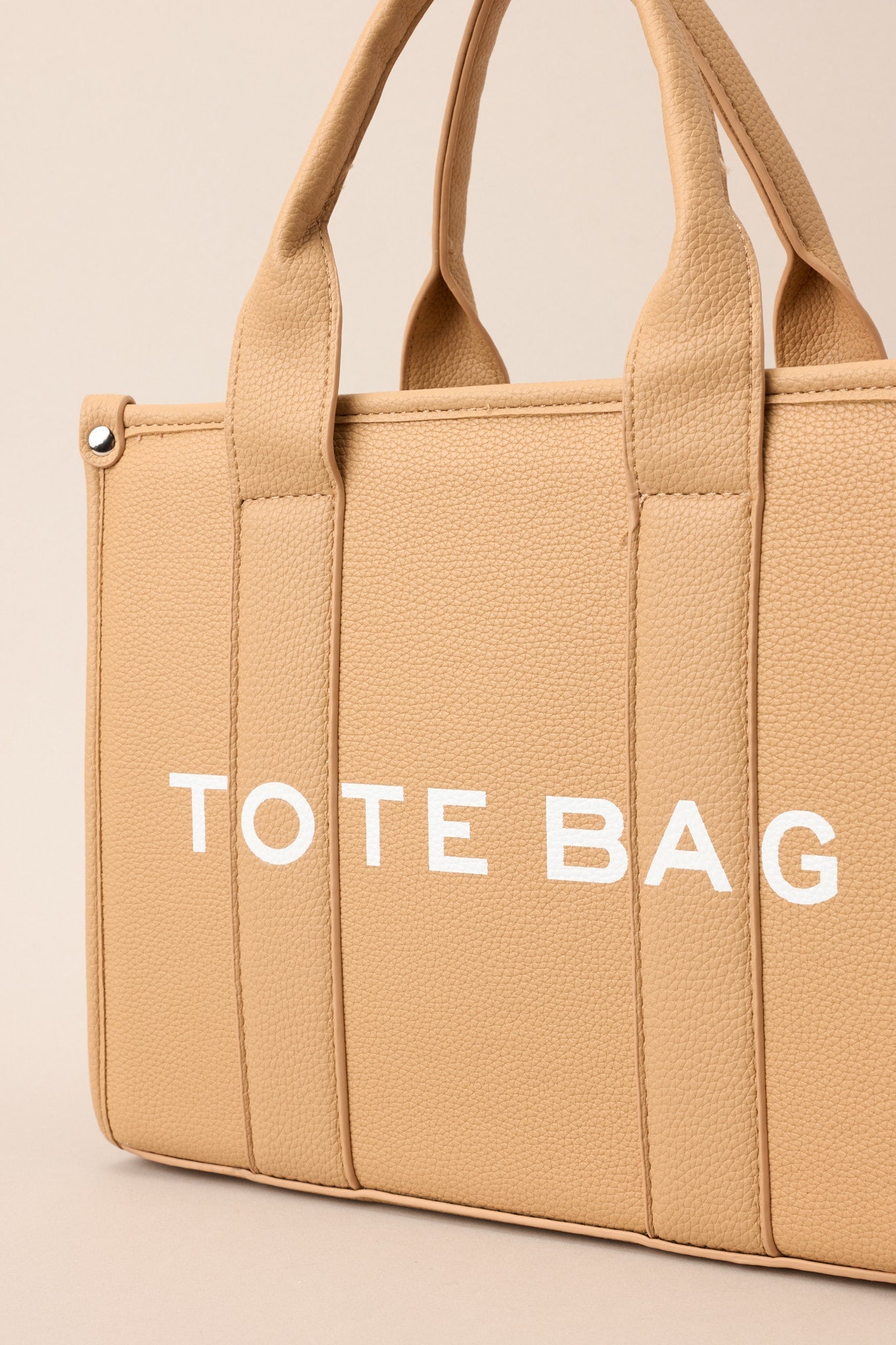 Close-up view of this tan bag that features silver hardware, top handles, a functional zipper closure, an additional pocket on the inside, and a removable strap.