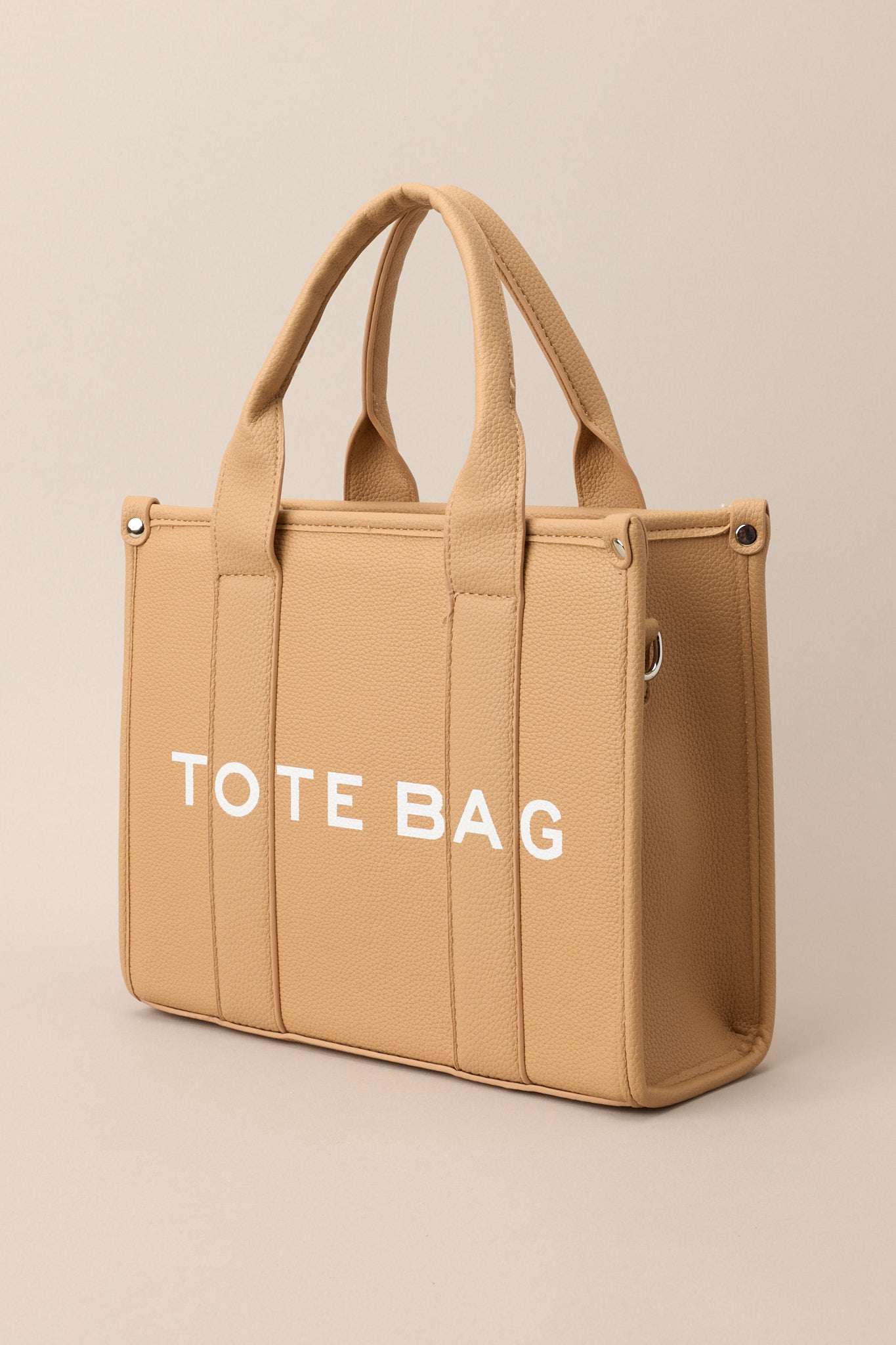 Side view of this tan bag that features silver hardware, top handles, a functional zipper closure, an additional pocket on the inside, and a removable strap.