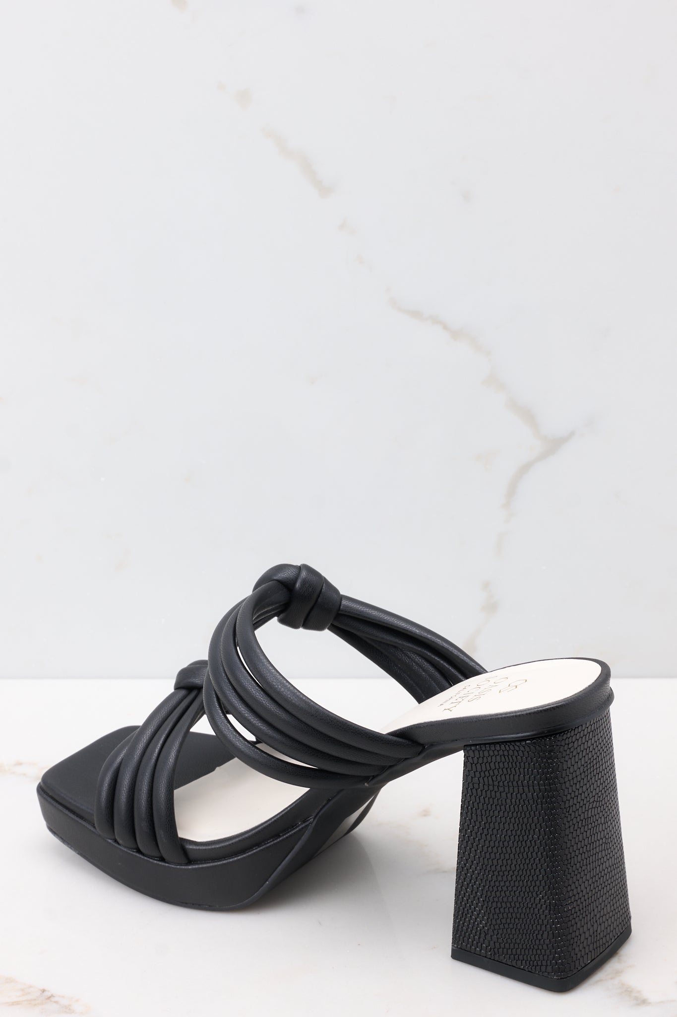 Back view of these black heels that feature a square toe, two straps across the top of the foot with knotted detailing, a small platform, and a block heel.