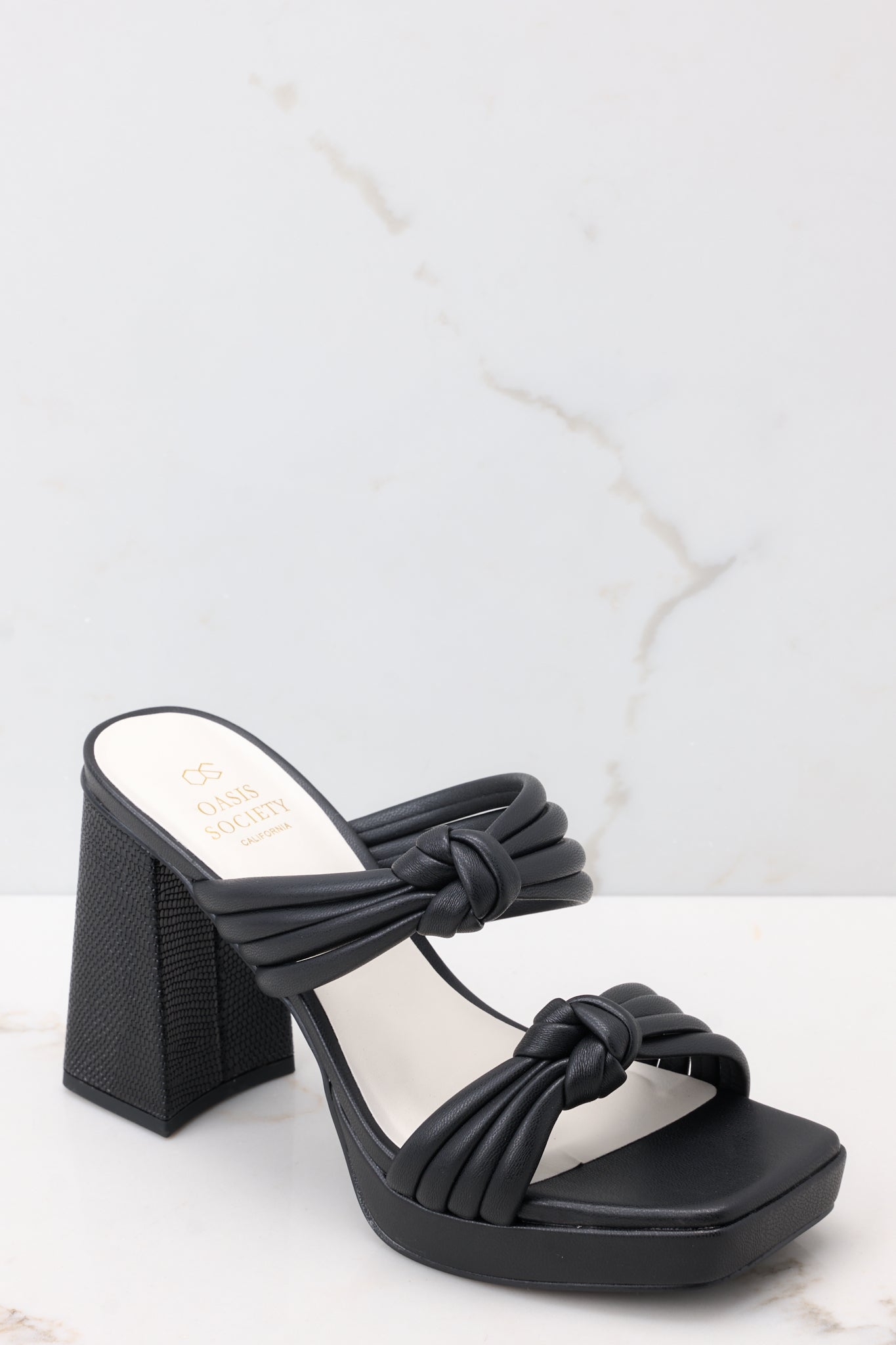 Angled view of these black heels that feature a square toe, two straps across the top of the foot with knotted detailing, a small platform, and a block heel.